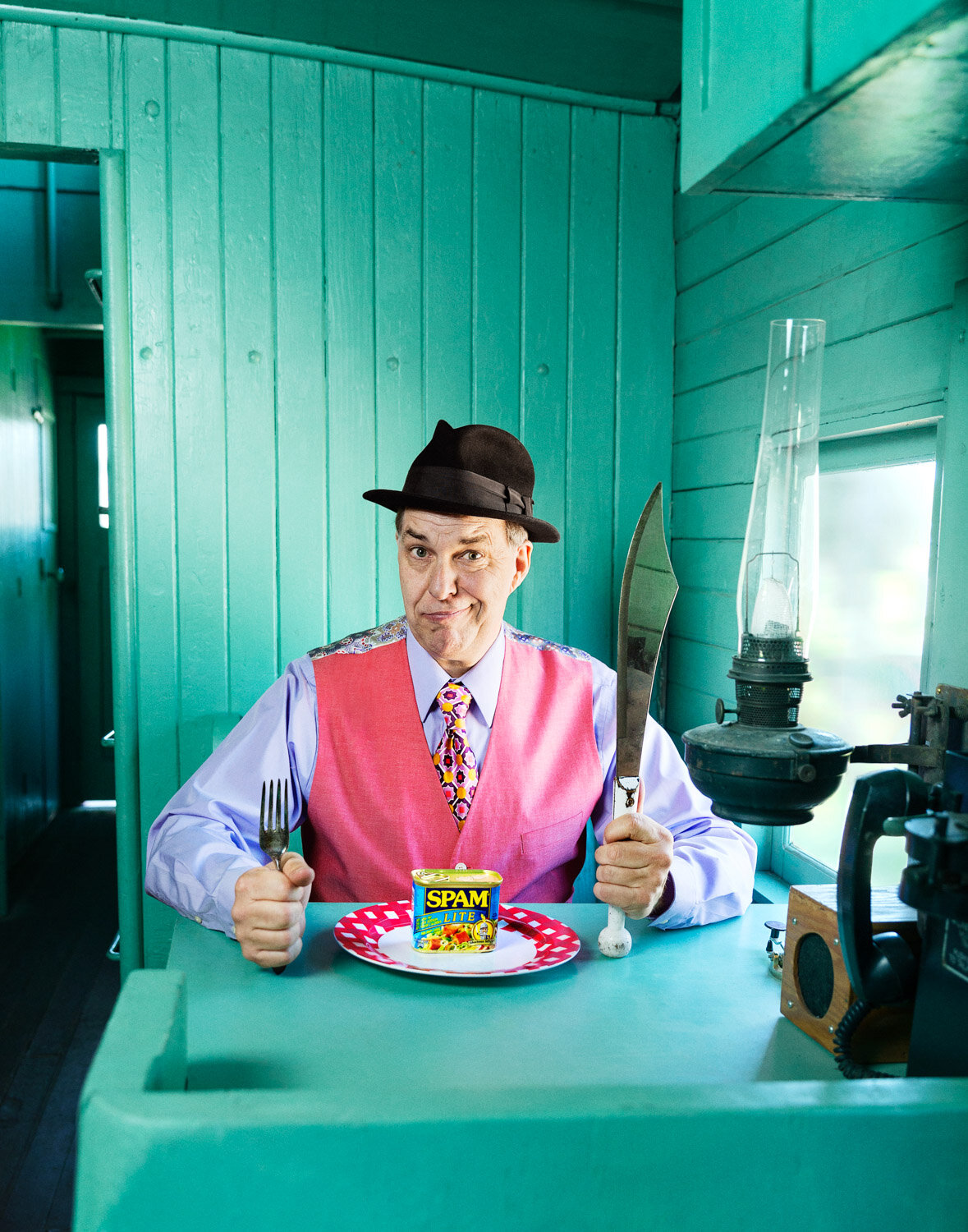 funny promotional portrait of juggler sitting in vintage train car with a machette, fork, and can of spam.