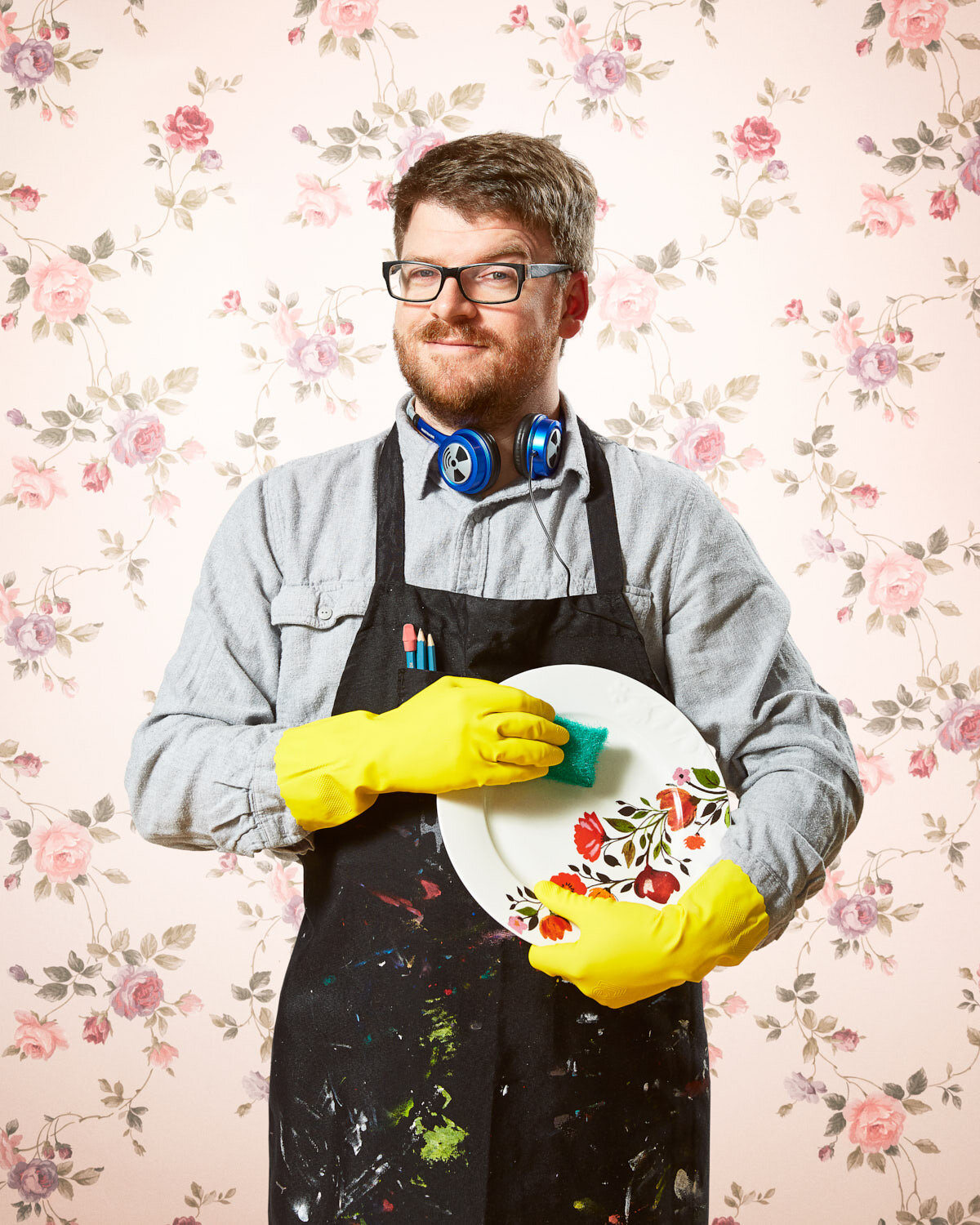 dad with headphones, artist apron, and dish gloves washing a dish with vintage floral wallpaper by conceptual portrait photographer Hanna Agar