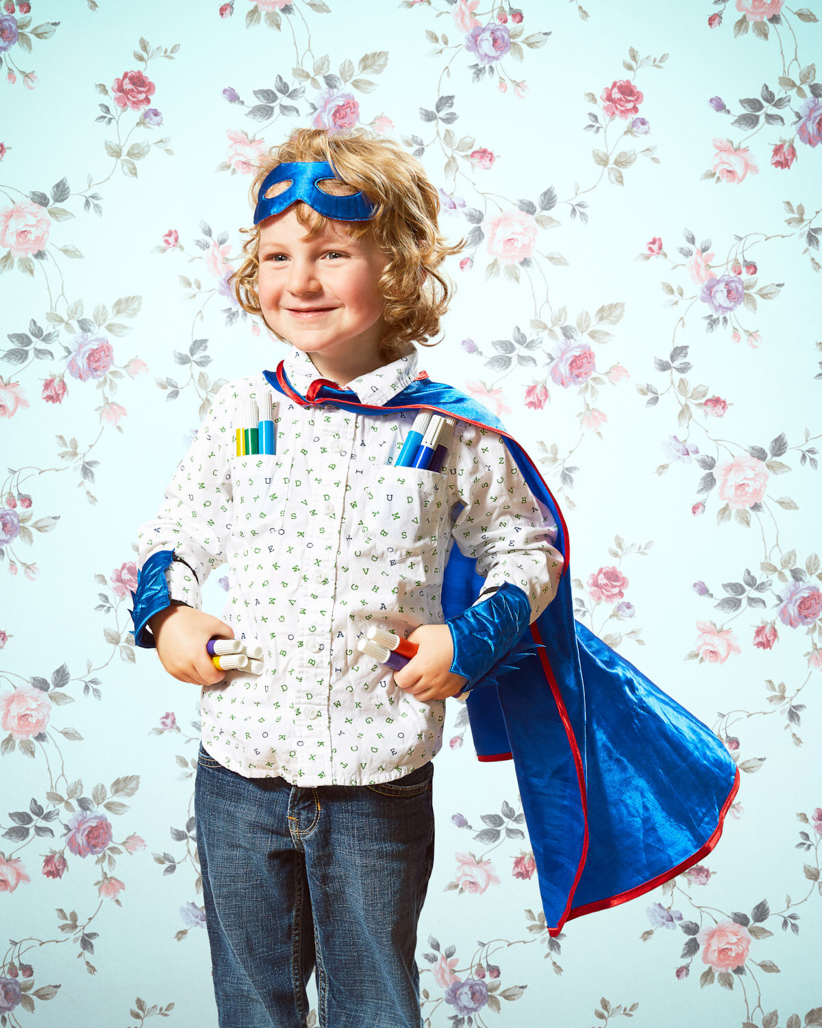 child wearing super hero cape and mask holding markers with vintage floral wallpaper by conceptual portrait photographer Hanna Agar