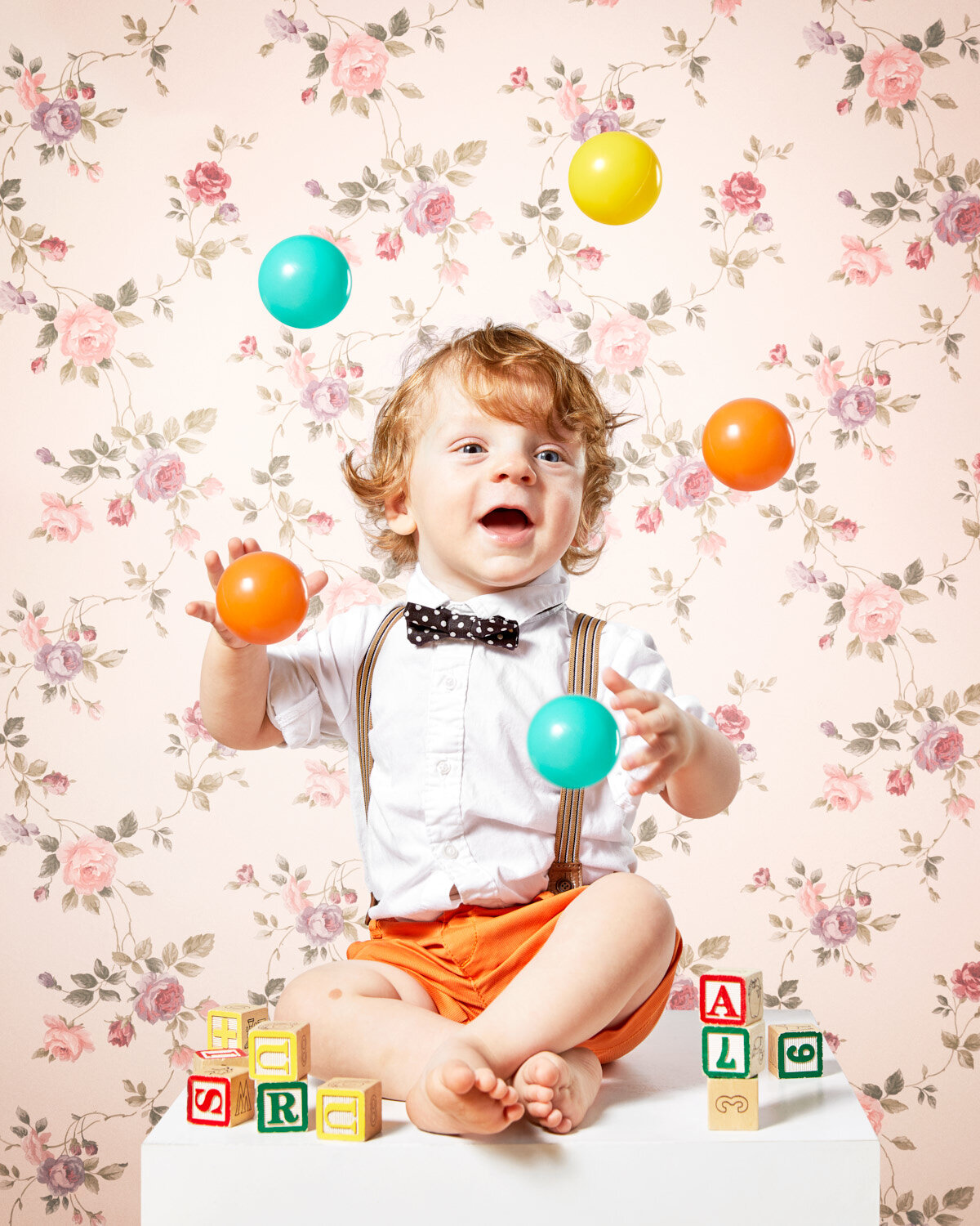 small child with bowtie and suspenders juggling colorful balls with vintage floral wallpaper by conceptual portrait photographer Hanna Agar