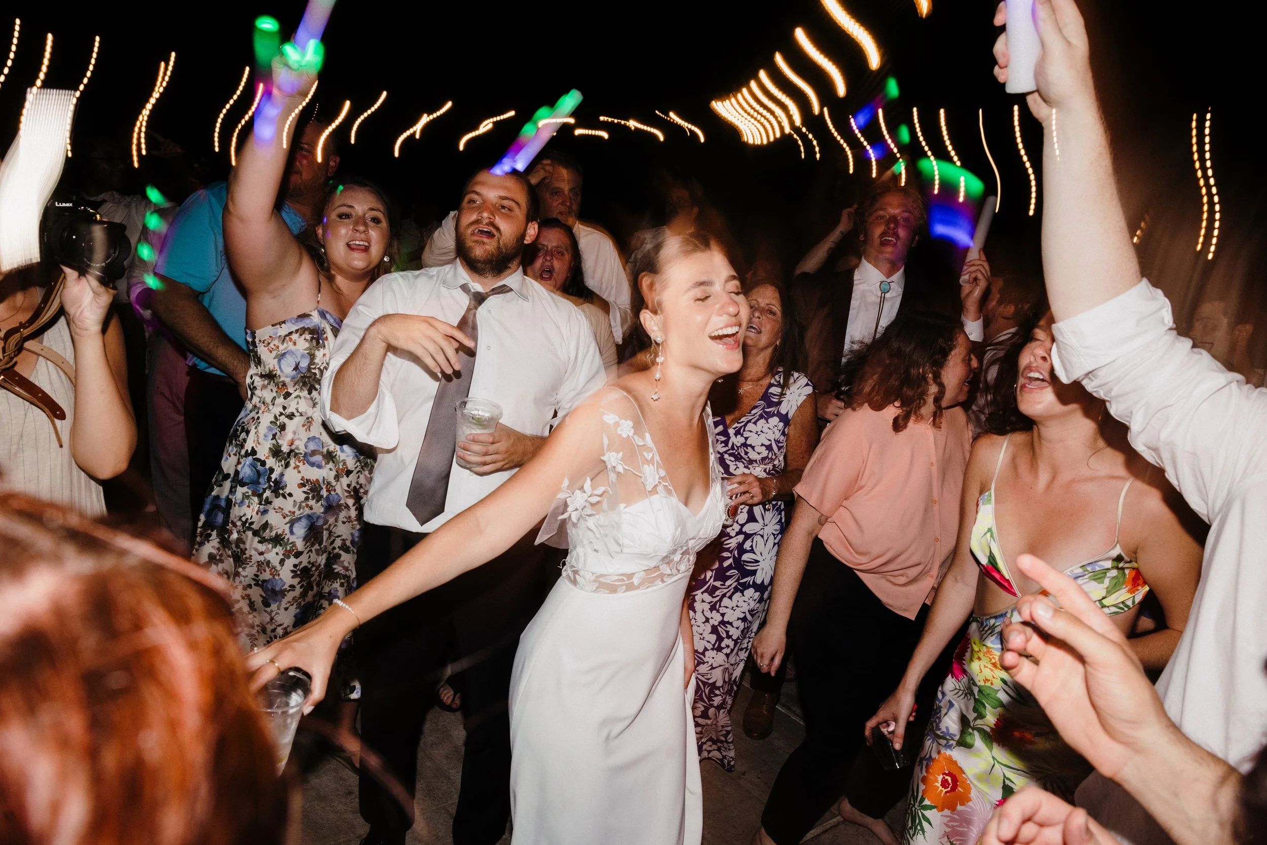 A joyful bride dancing at her wedding reception with guests around her, some singing and others smiling, under colorful blurred lights at night.