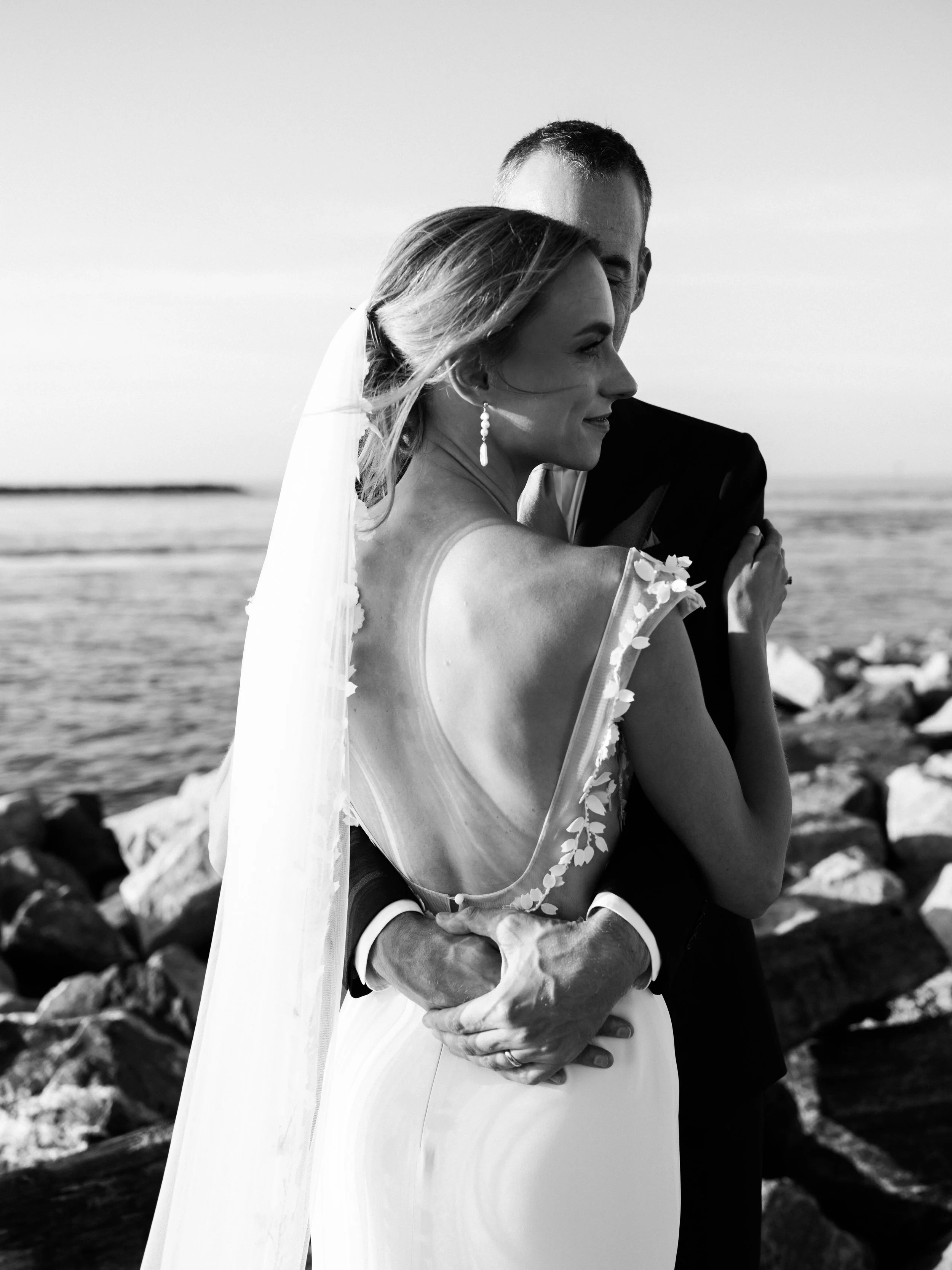 A bride and groom embracing on a rocky beach, with the ocean in the background, during their wedding. The bride has a veil and a dress with floral details, and the groom is in a tuxedo.