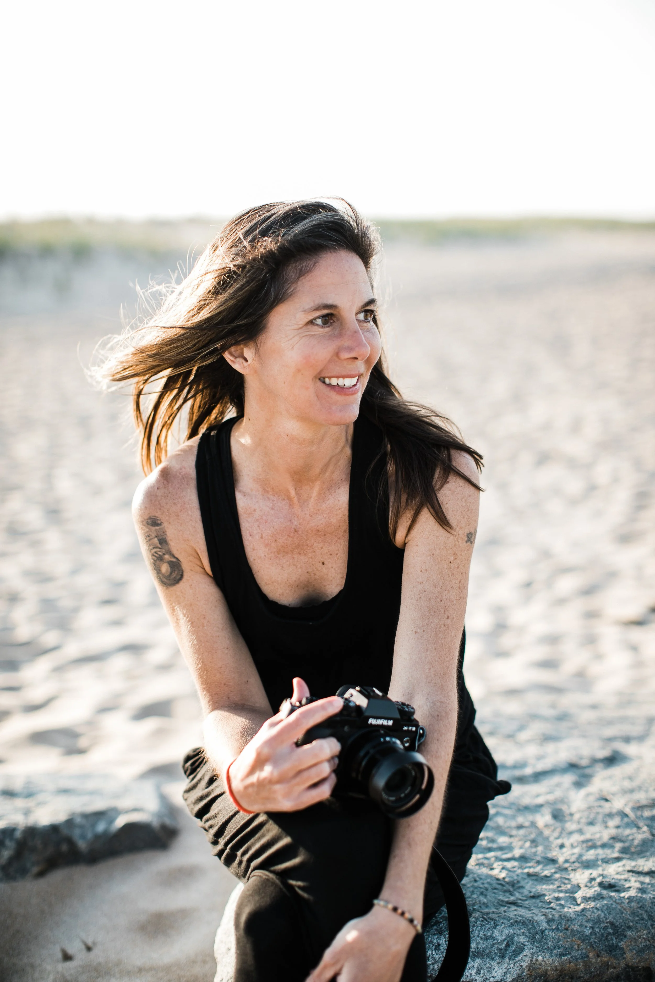 A woman with long brown hair, wearing a black tank top, is sitting on a rock on a sandy beach, holding a camera, smiling and looking to her right, with a bright sky in the background.