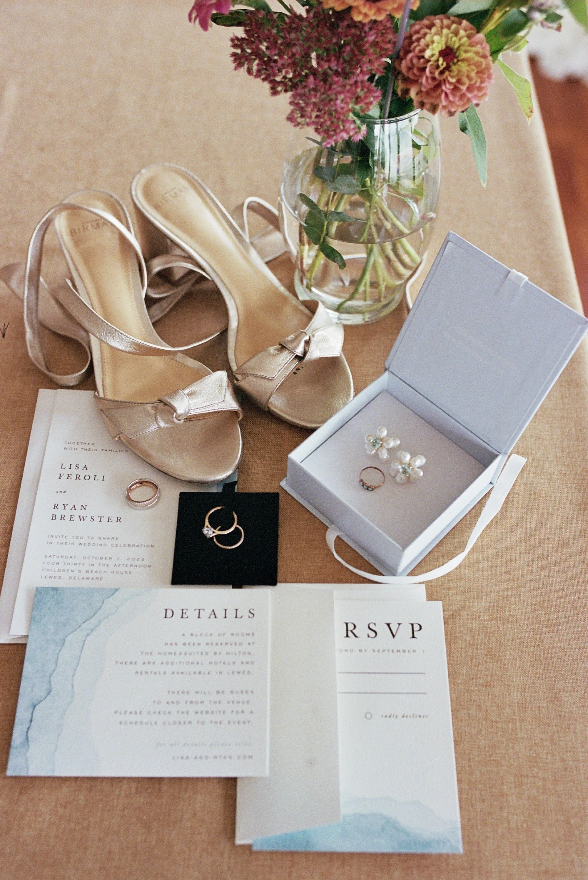 A wedding invitation, gold high-heeled shoes, a small ring box with pearl earrings, a ring, and an RSVP card arranged on a table next to a vase of pink and burgundy flowers.