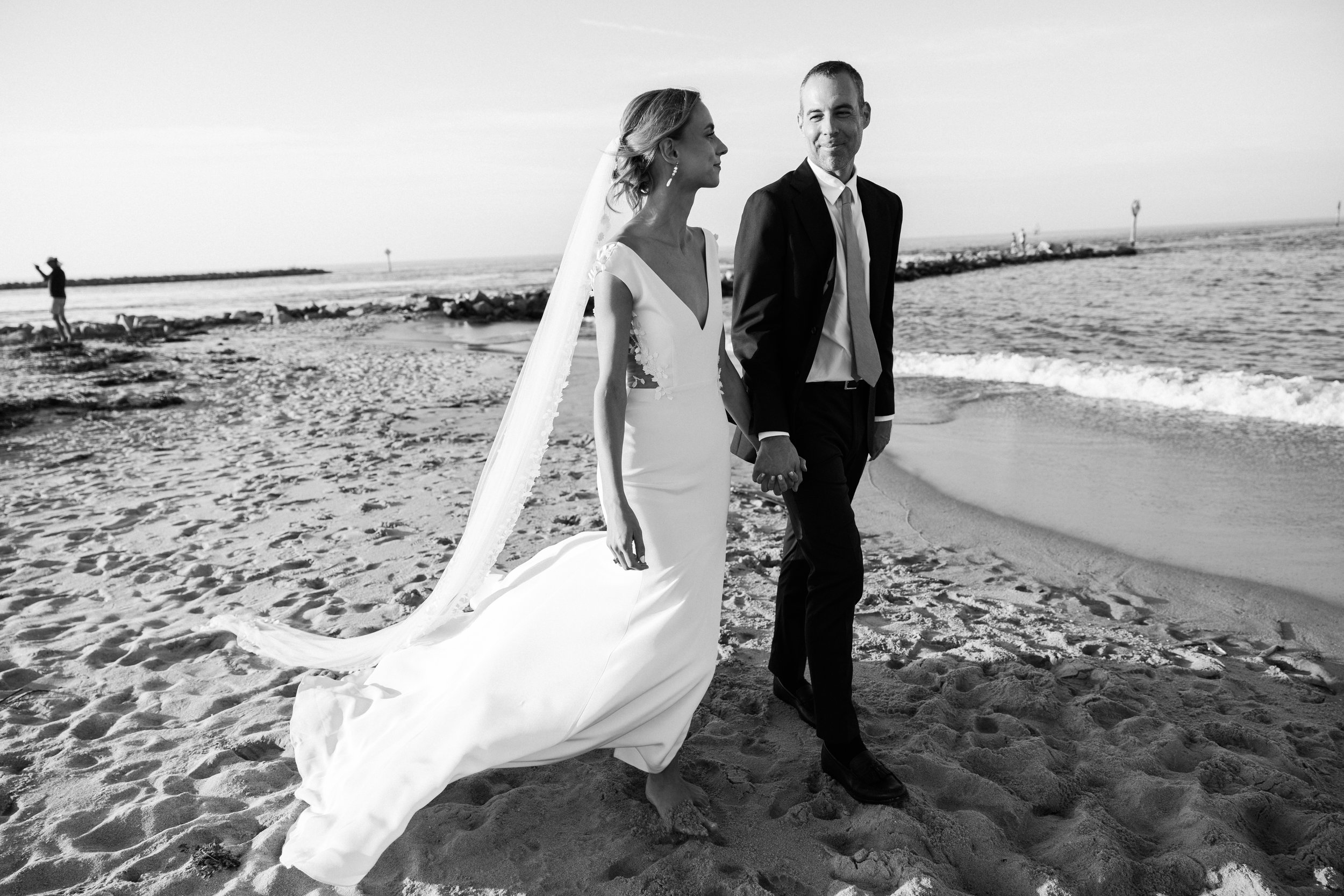 Black and white photograph of a bride and groom walking hand in hand on a sandy beach, with the ocean and a man taking a photo in the background.