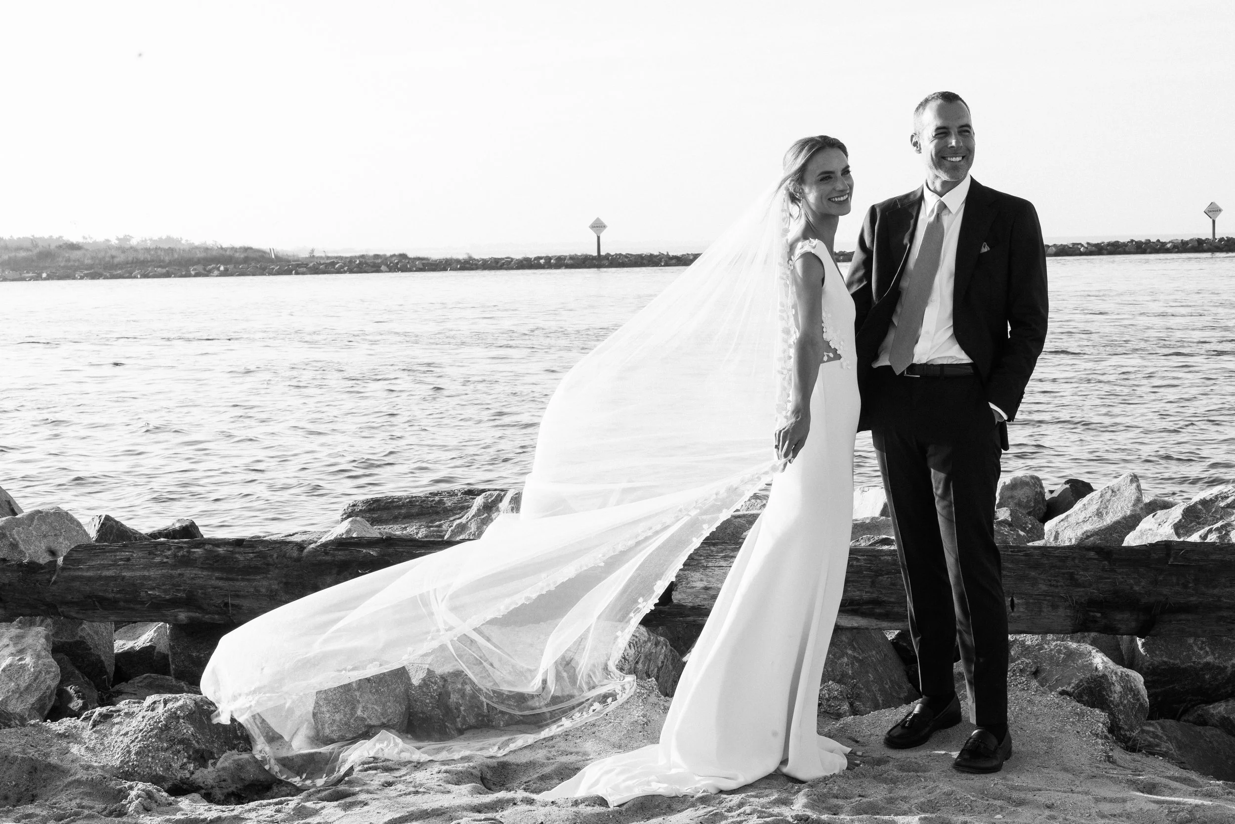 A bride and groom in wedding attire standing on a rocky shoreline by the water, smiling and facing the camera.