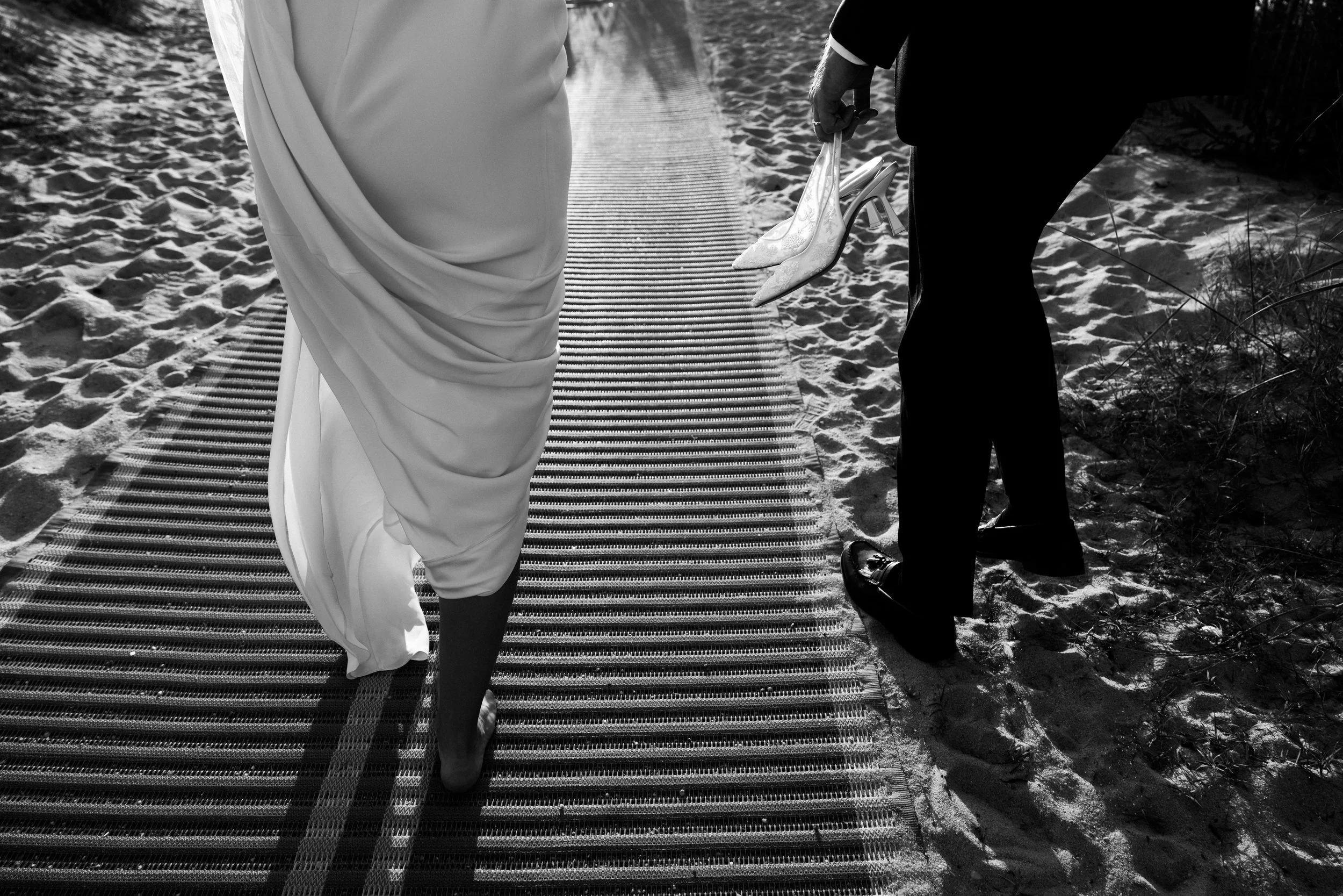 A person in a white dress walking on a sandy beach next to another person dressed in black, holding a shoe, with a beach mat on the sand.