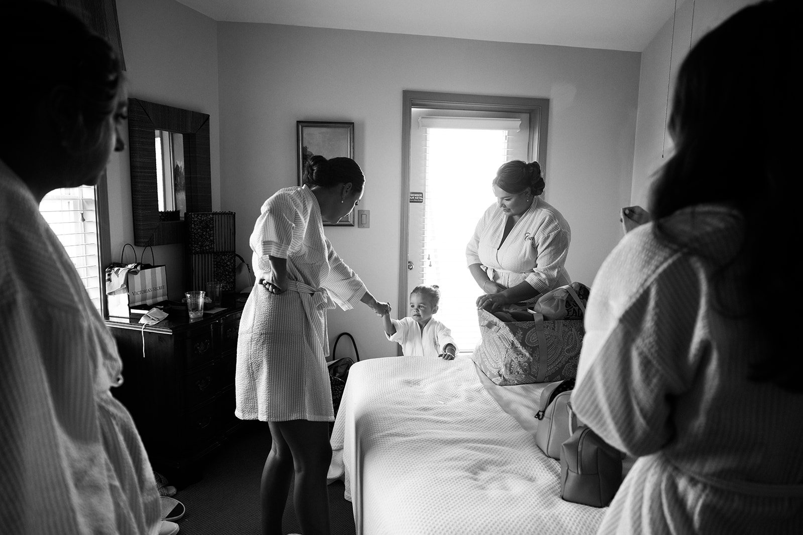 Bridesmaids getting ready, holding hands in a bright room with bags and belongings, as part of a visit or welcoming moment.