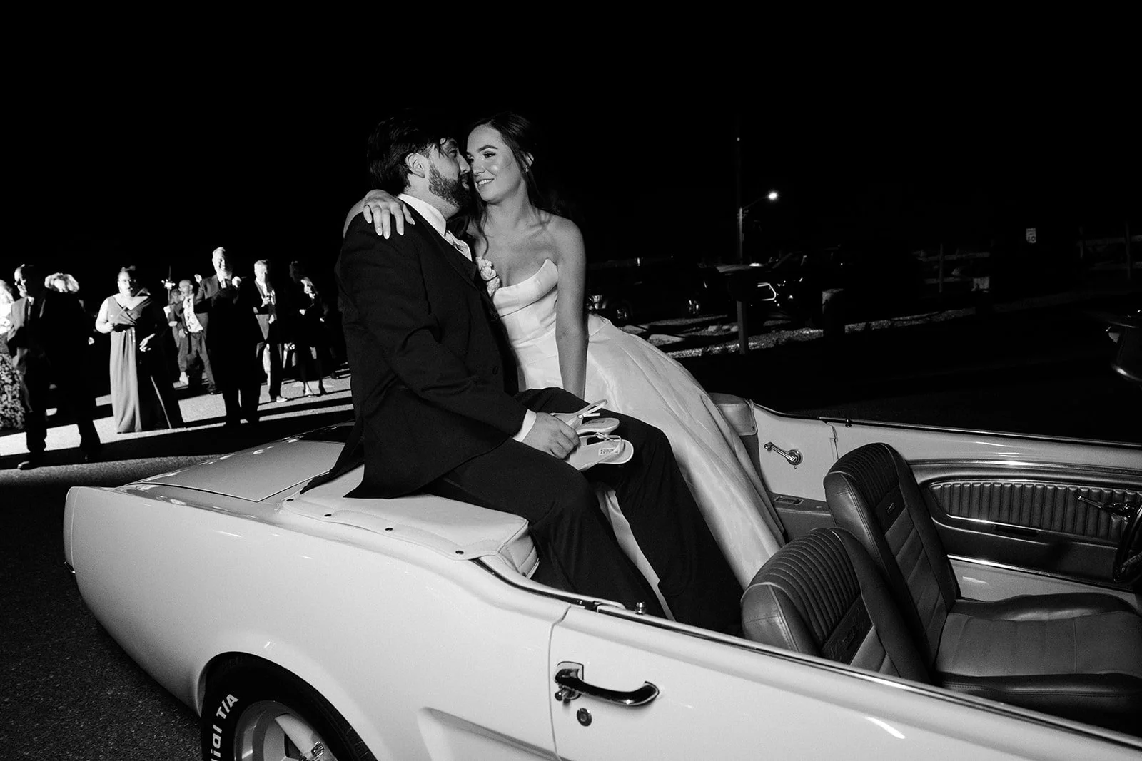 A black and white photo of a newlywed couple sitting on the back of a vintage convertible car at night. The groom is dressed in a suit, and the bride is wearing a wedding gown. They are smiling and looking at each other, with wedding guests standing 
