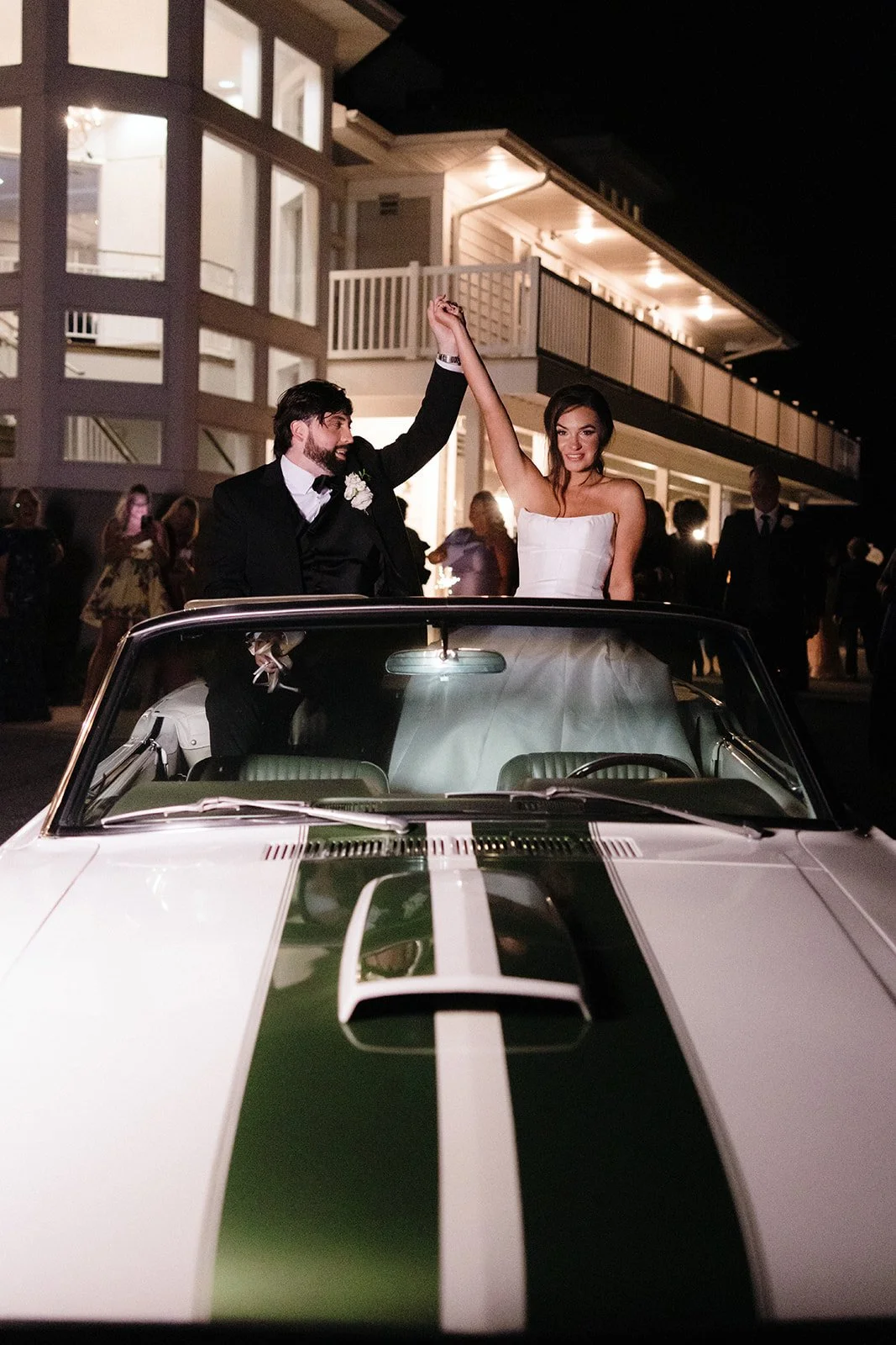 A newlywed couple celebrating in a vintage white convertible car at night. The groom is in a black tuxedo and the bride is in a white wedding dress, holding hands and smiling. There are guests in the background outside a lit house.