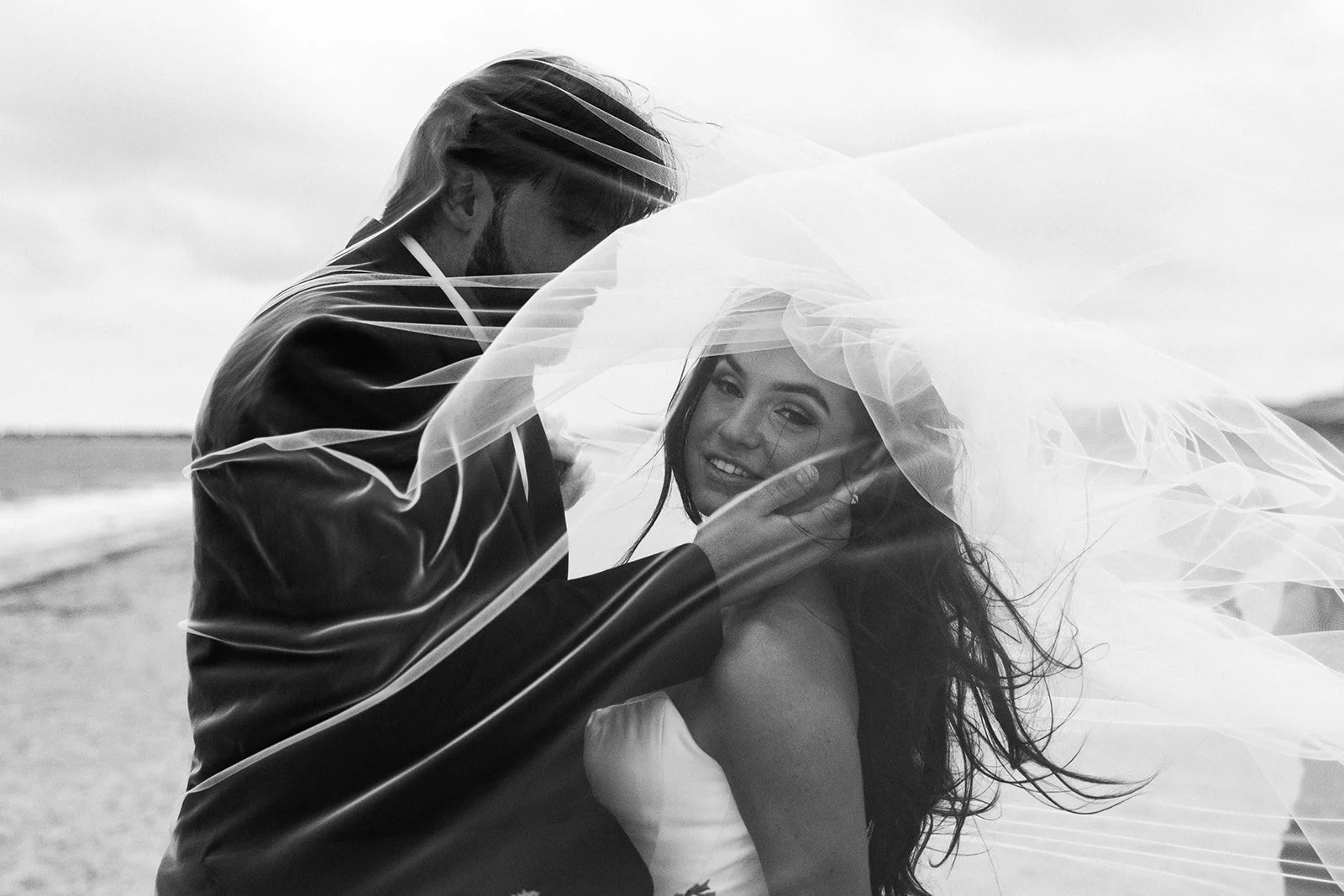 A black and white photo of a bride and groom on the beach, with the groom leaning over the bride, who is smiling under her veil.