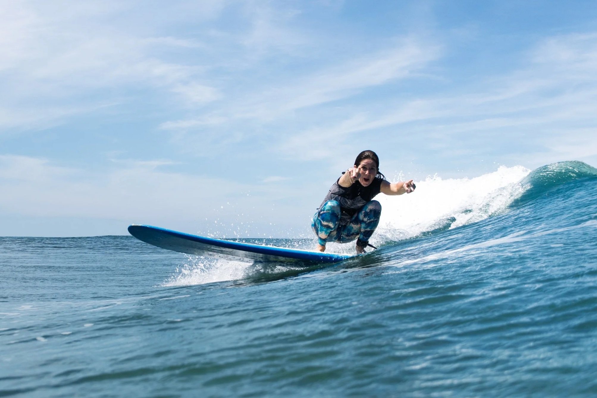 Woman surfing on a blue surfboard in the ocean, pointing forward with an excited expression, under a partly cloudy sky.