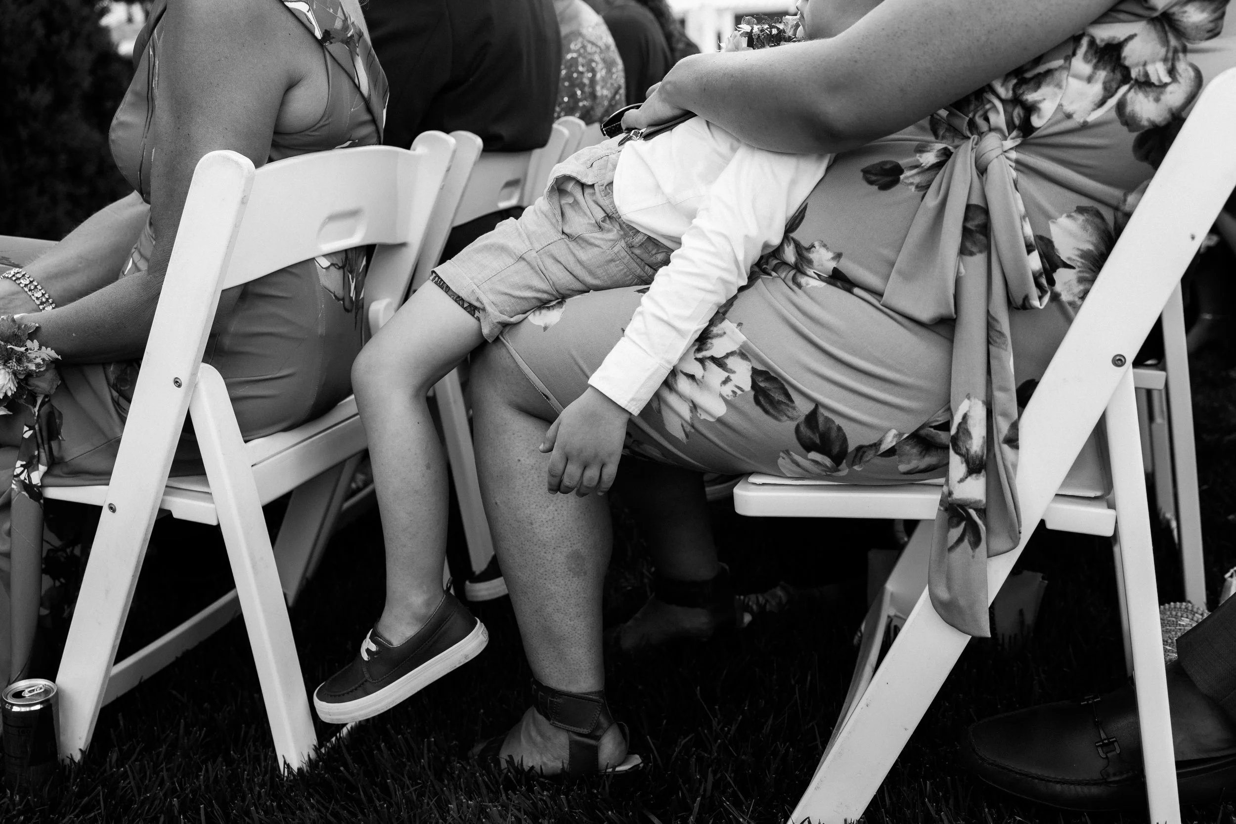 People sitting on white chairs outdoors, focusing on a woman wearing a floral dress and a young boy in shorts and a long-sleeve shirt, with legs crossed and hands resting on her lap.
