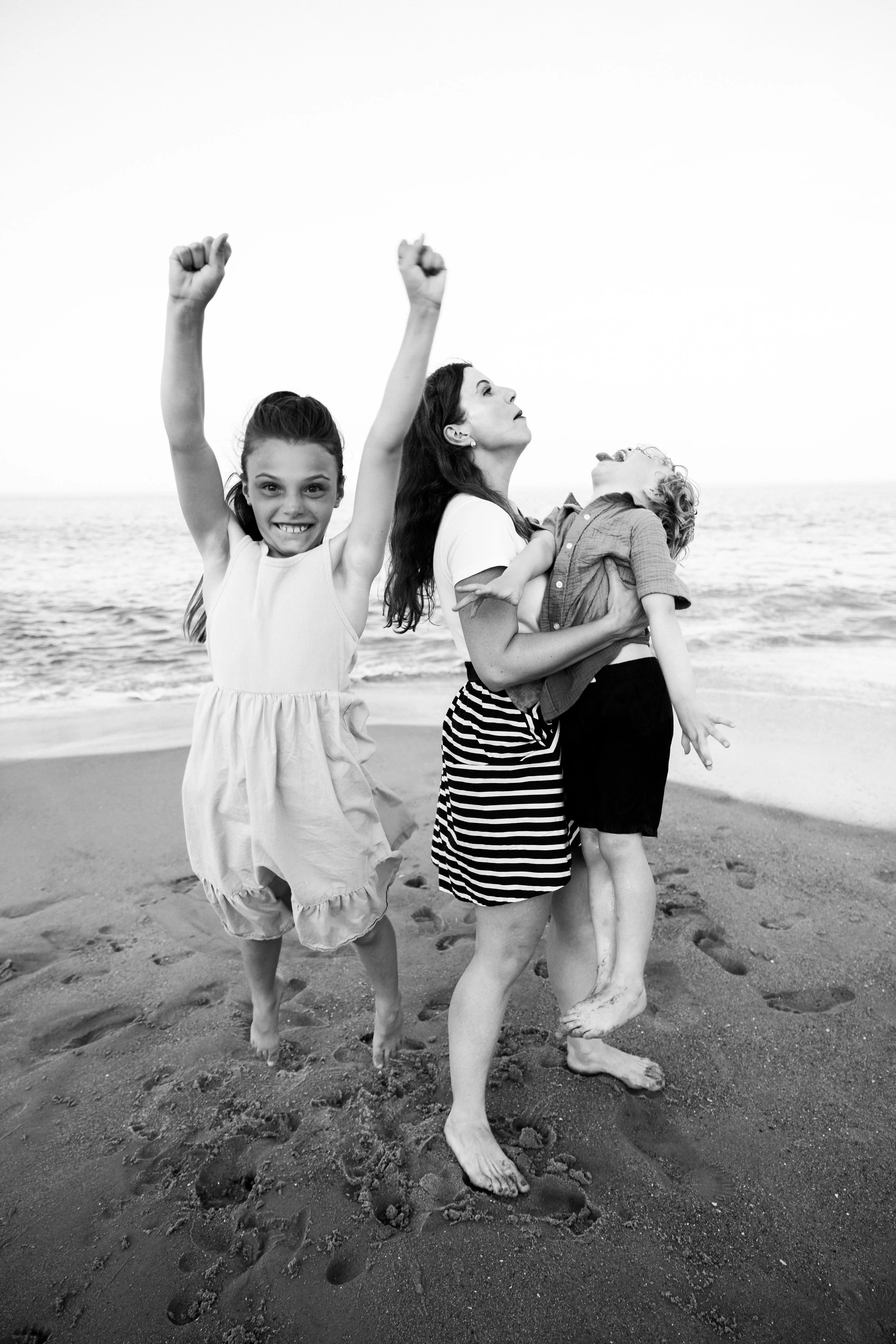 A woman and two children enjoying a day at the beach, with the woman holding a child in her arms and the girl nearby with her arms raised in excitement.