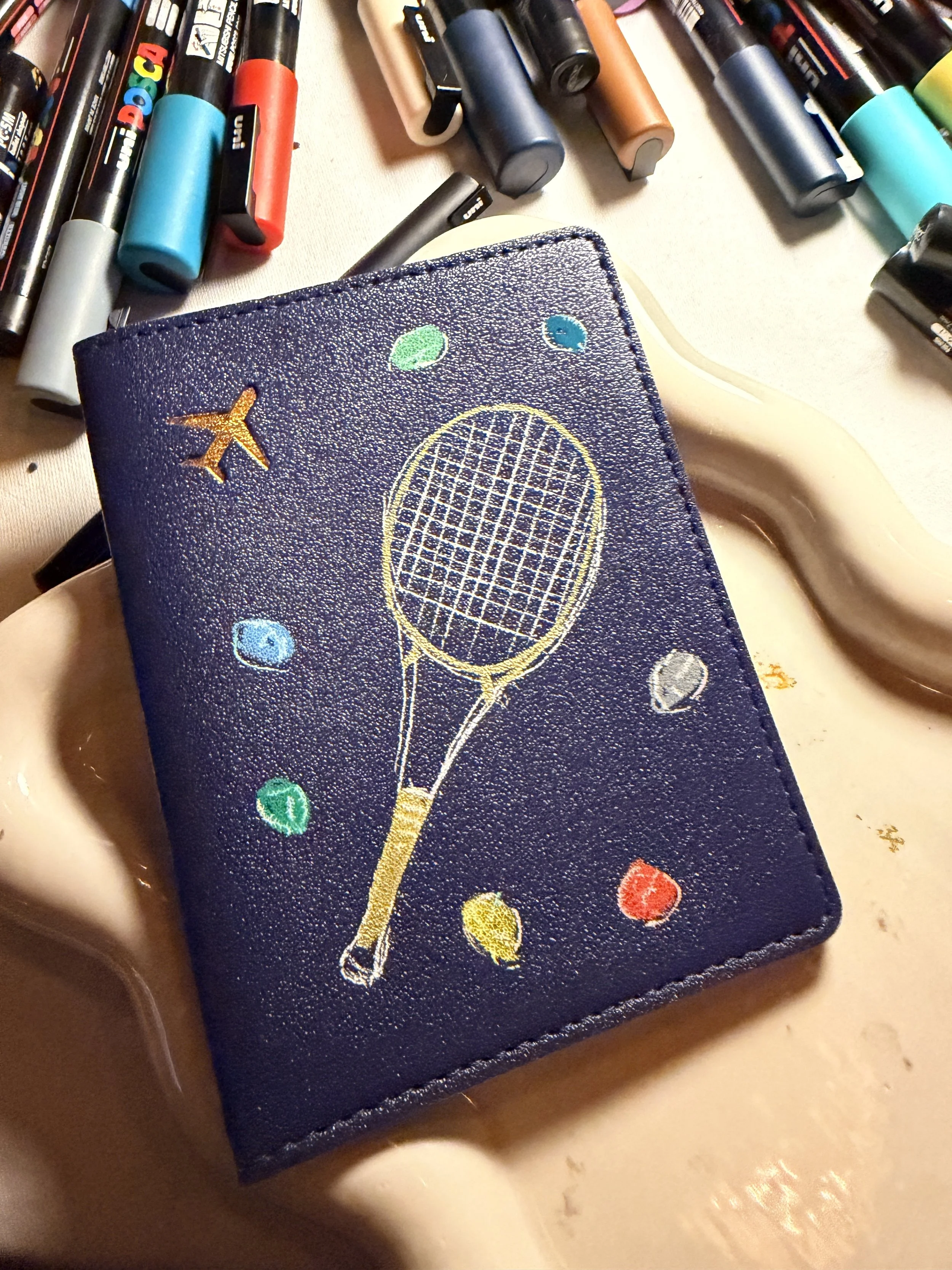 Custom hand-painted navy passport holder with a gold tennis racket and colorful gemstone dot illustration — live brand activation by Danielle Rose Fisher NYC