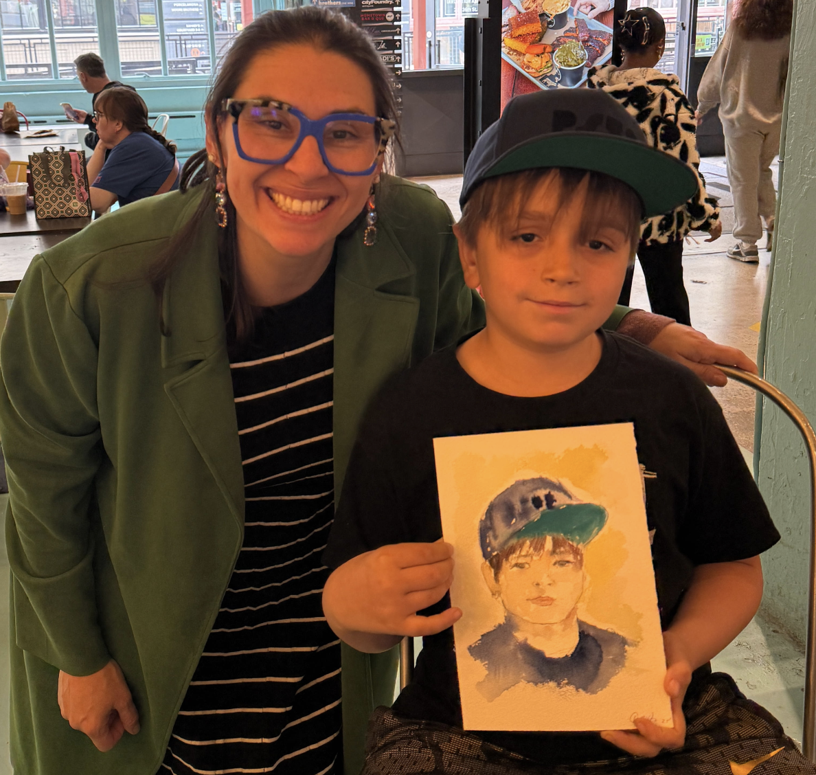Danielle Rose Fisher with a young guest holding his finished live watercolor portrait at an Industry City event — the boy wearing a baseball cap, matching his illustration