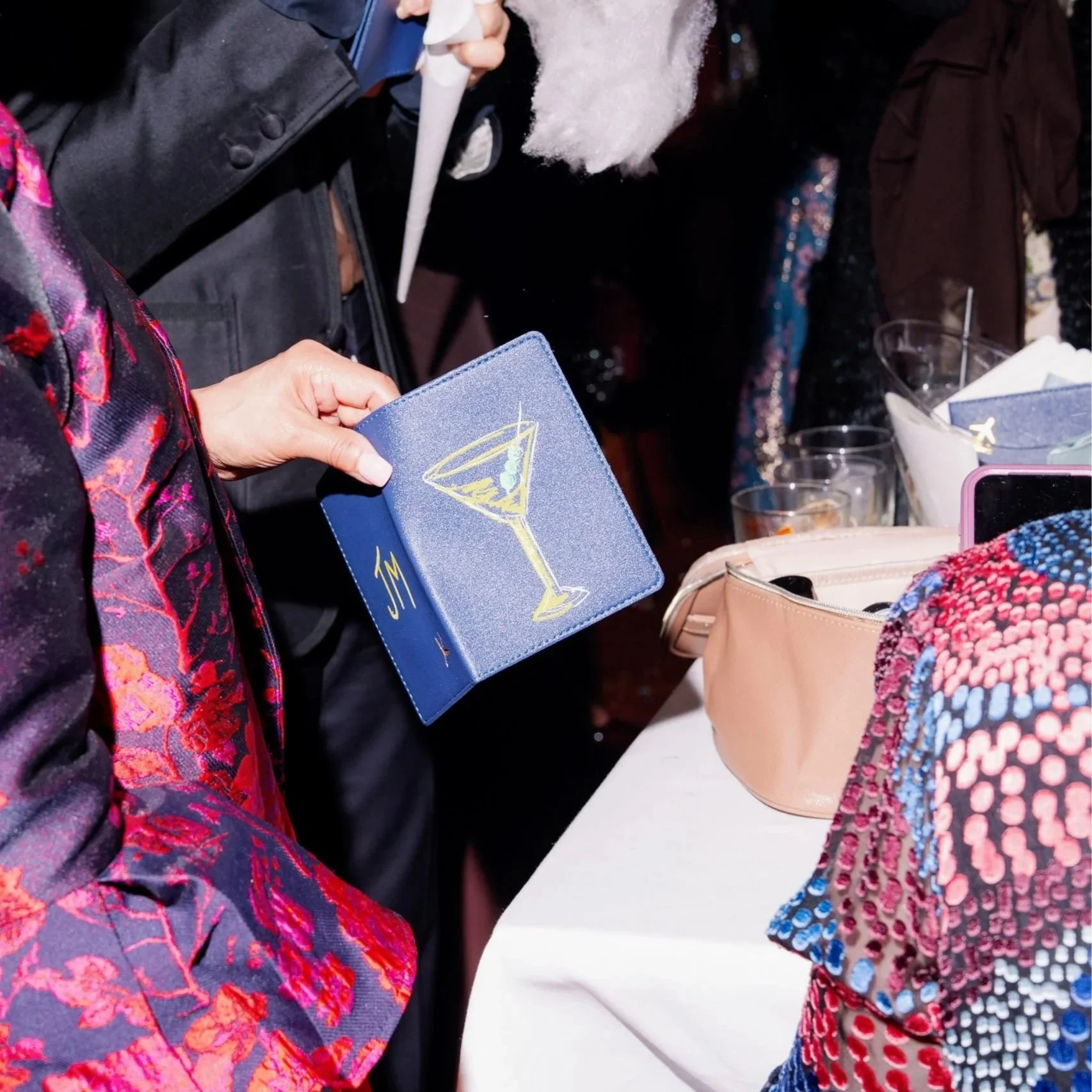 A person holding a blue card with a drawing of a martini glass and the initials 'JM' in gold design, with another person in the background attempting to spray a white foam or mist into their face.