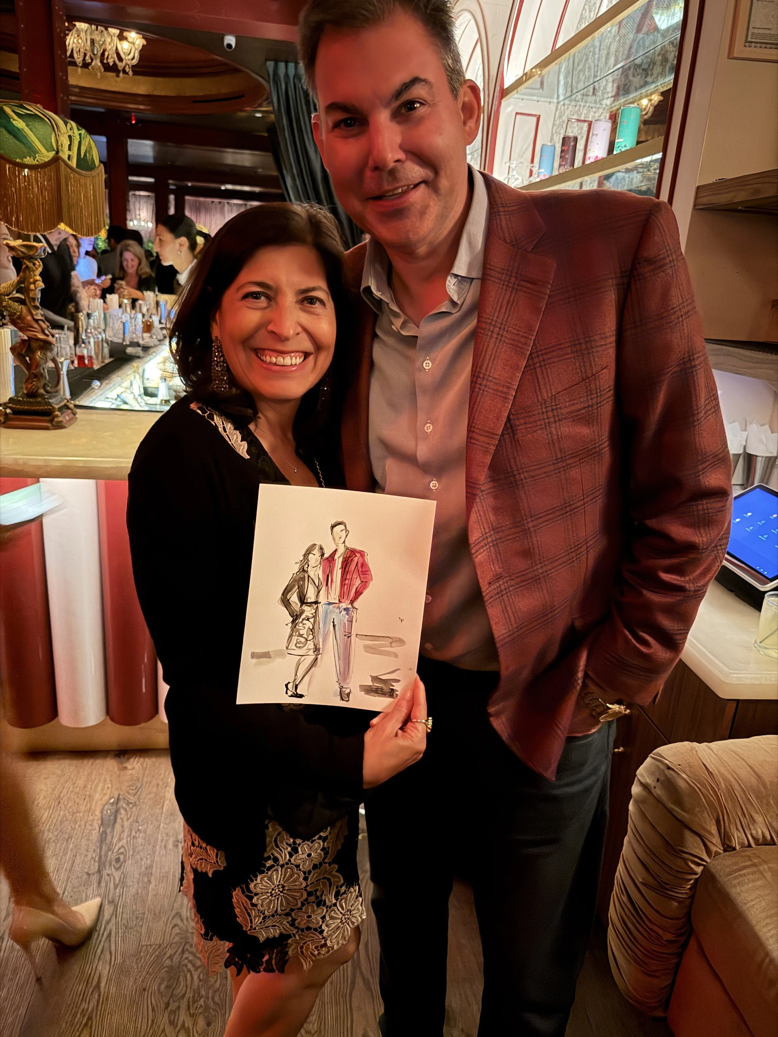 Two guests at the Angel Ball Launch Party holding their live fashion illustration by Danielle Rose Fisher — figures in red and lavender tones