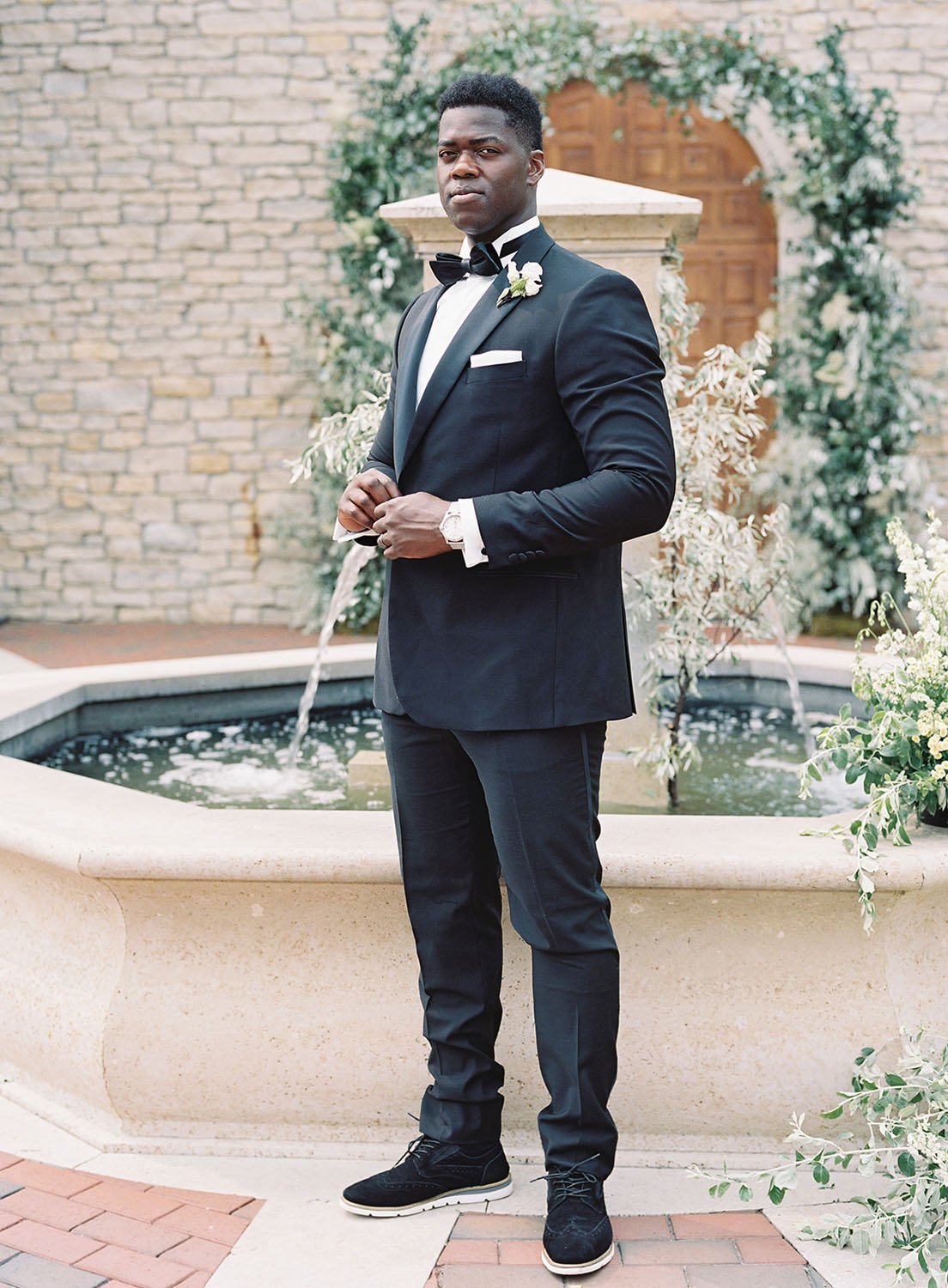  a groom wearing a black tux standing in front of a fountain outside of a villa in Wisconsin 