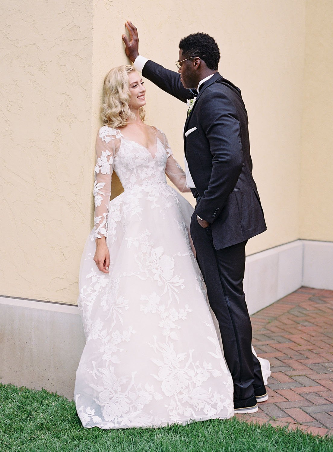  A bride and groom leaning against a wall in a courtyard during their spring garden wedding in Wisconsin. The bride is wearing 'Maeve' by Monique Lhuillier wedding dress. 