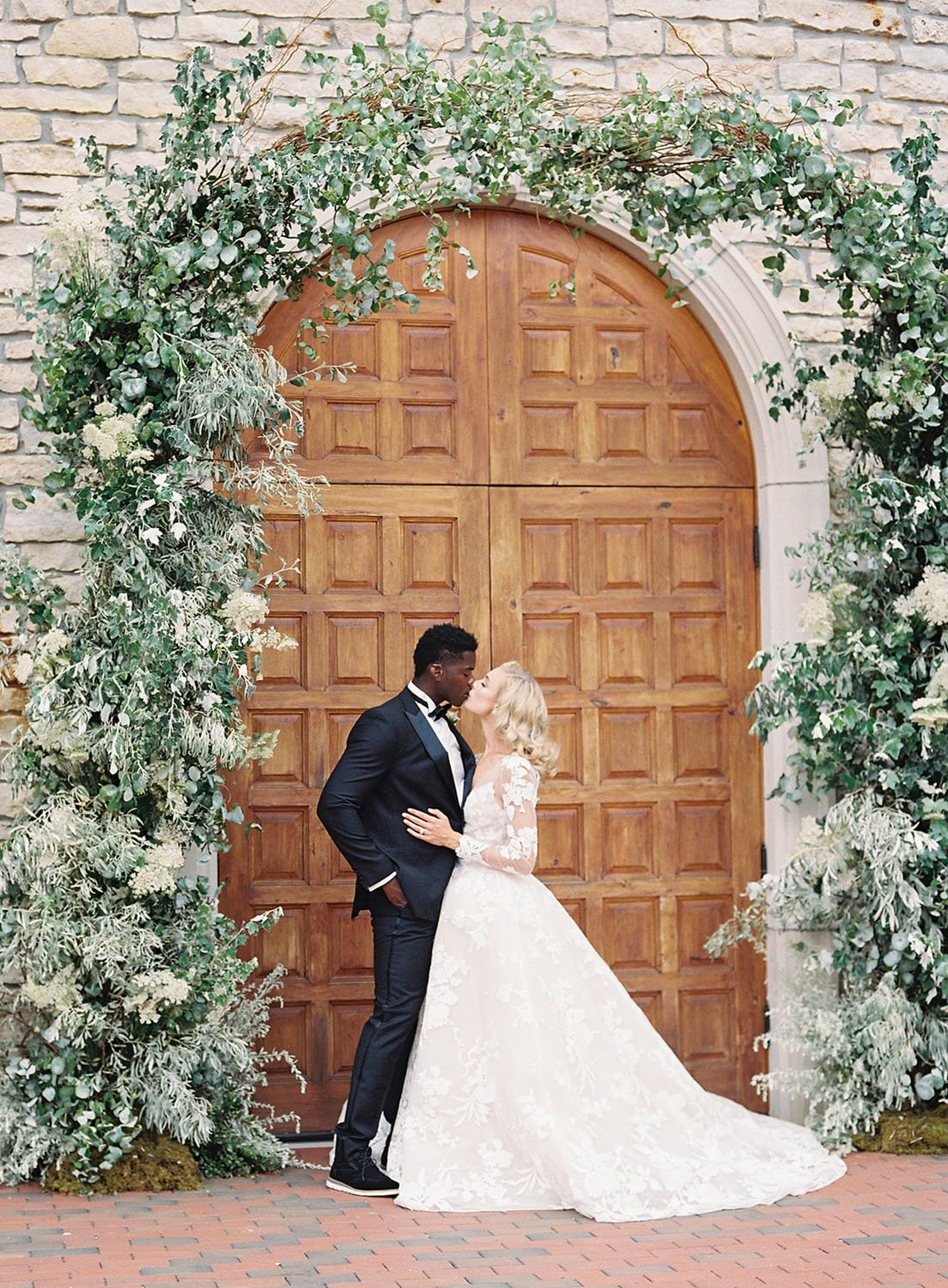  A bride and groom sharing a kiss under a greenery arbor for their spring midwest wedding. The bride is wearing 'Maeve' by Monique Lhuillier. 