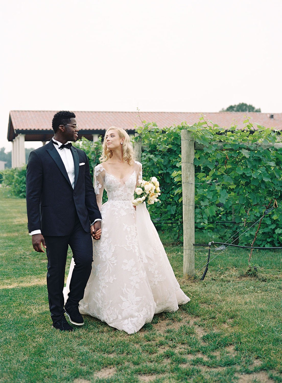  A bride and groom holding hands and walking through a vineyard garden for their spring midwest wedding. The bride is wearing 'Maeve' by Monique Lhuillier wedding gown. 
