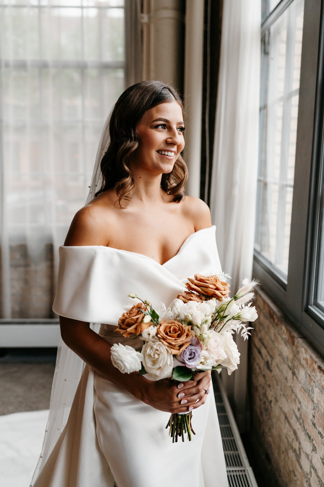 the happy bride looking out the window in her off the shoulder wedding dress by eva lendel