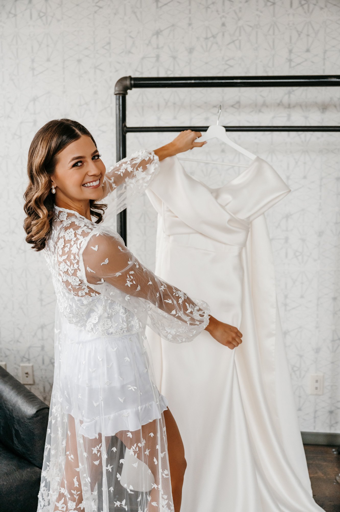 bride getting ready in a floral white lace bridal robe looking at her wedding dress hanging in a modern setting