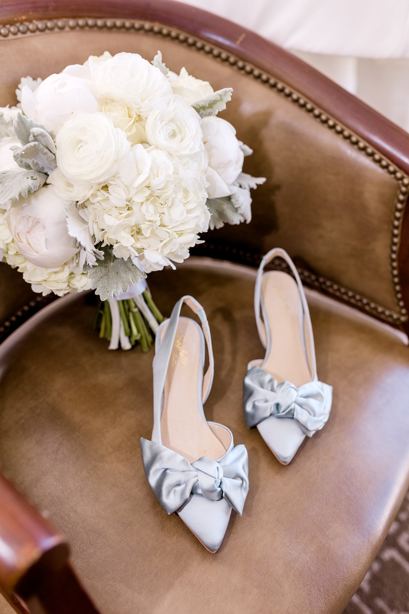  Classic bridal bouquet with white peonies, hydrangea, ranunculus, and lambs ear propped in a brown leather chair with silver bridal shoes with a pointed toe and large bow 