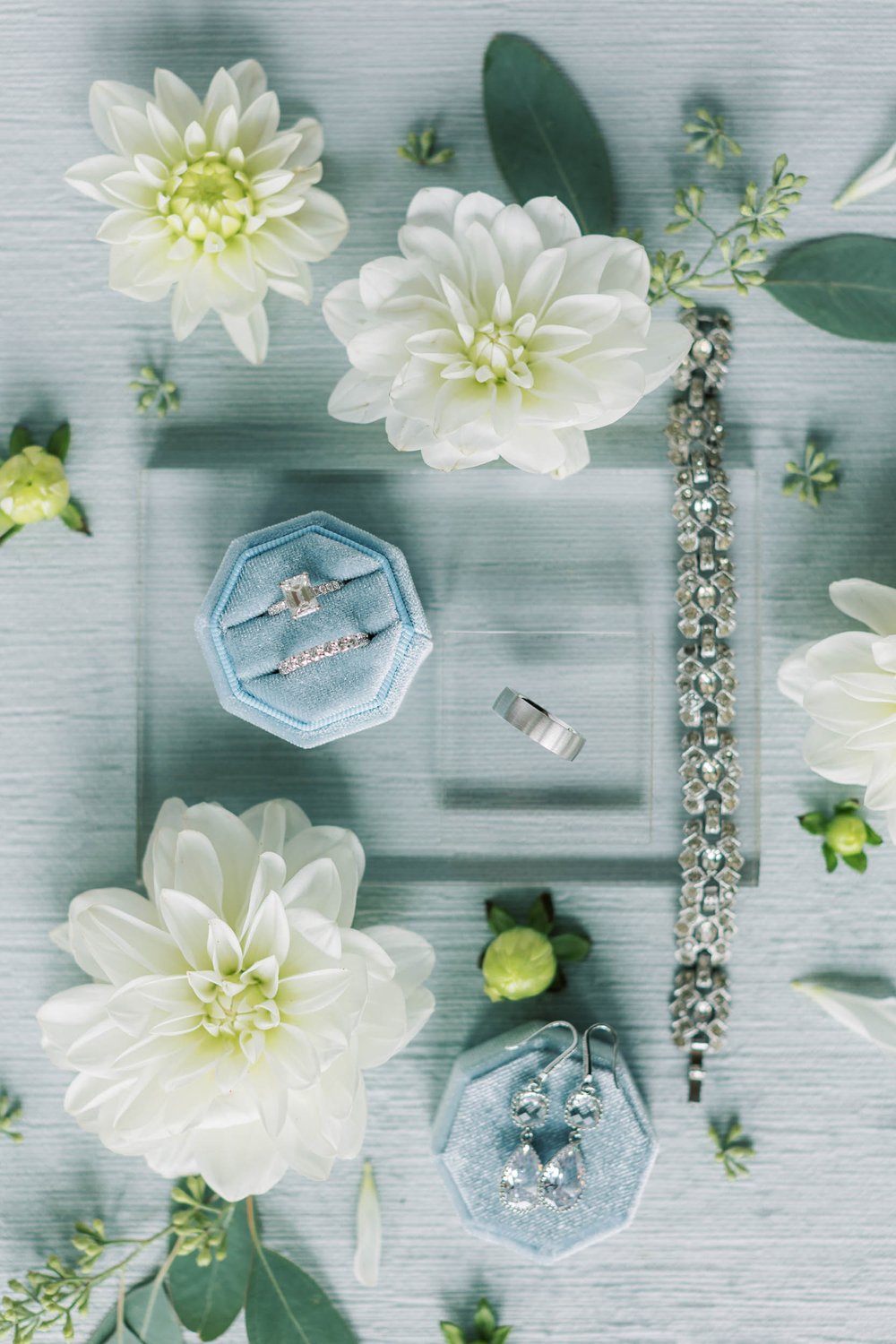  a beautiful wedding flat lay on a blue background featuring the couples’ wedding jewelry, rings, florals, and other sentimental items for their big day 
