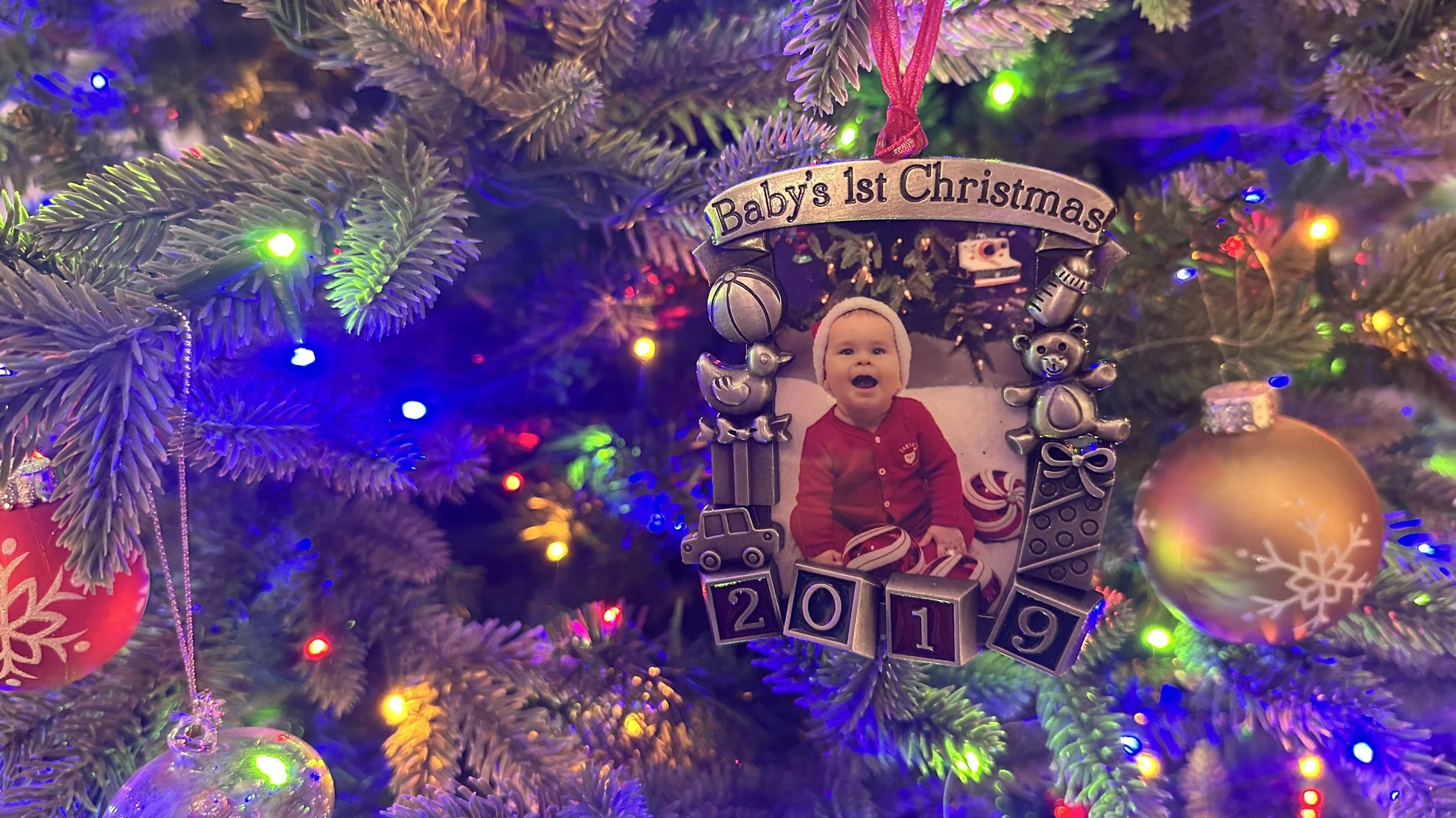 A Baby’s First Christmas ornament from 2019 hanging on our Christmas tree, surrounded by lights and ornaments.