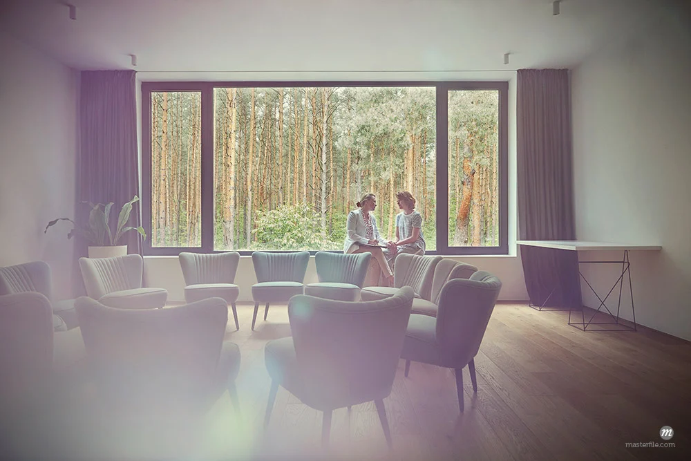 Women talking at window in group therapy room © Masterfile - Person(s) appearing in the above photograph are models and used for illustrative purposes only.