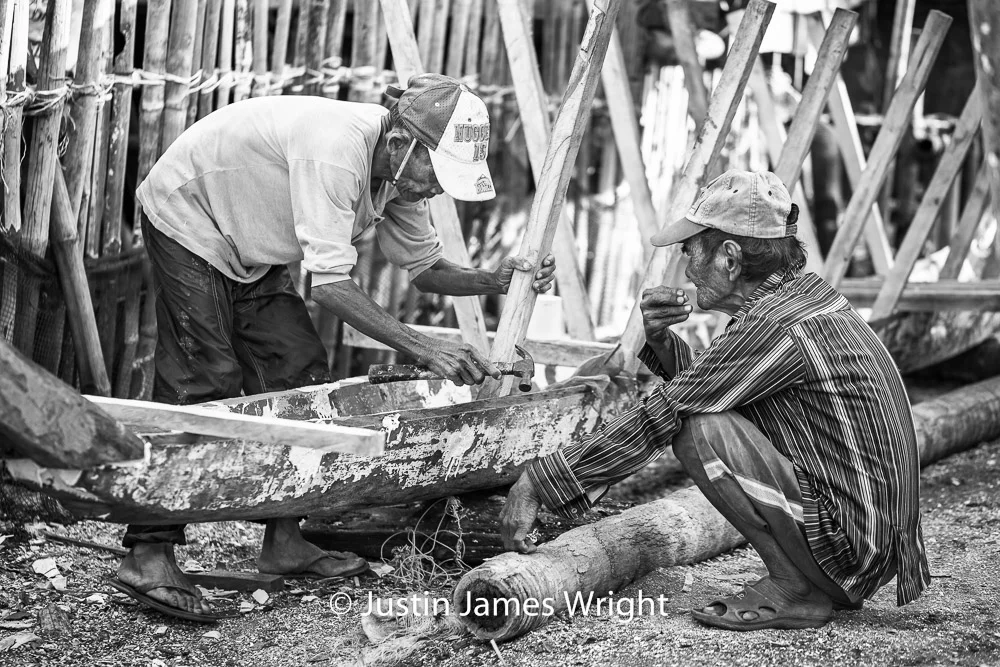 The Boat Builder Mang Alog enacts the art of crafting a banca to a captive audience of one; tobacco-chewing Mang Aleo. Resilience - The Isla Pulo Community, Philippines. January 2013. Canon EOS Mk III, EF 100 mm, F 2.8, 1/400 sec.