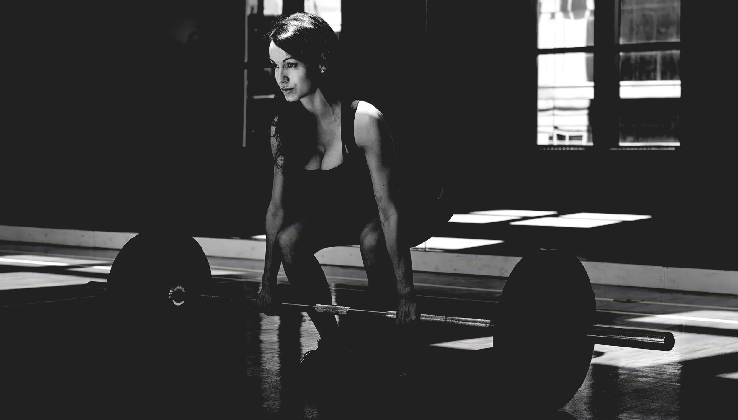 Black-and-white photograph of a woman preparing to lift a loaded barbell during strength training.