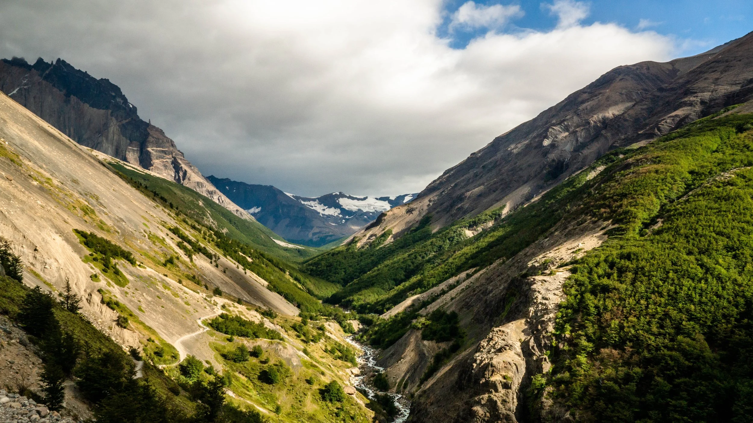 A scenic mountain valley with steep, rocky slopes on either side, lush green vegetation at the bottom, and snow-capped peaks in the distance under a cloudy sky.