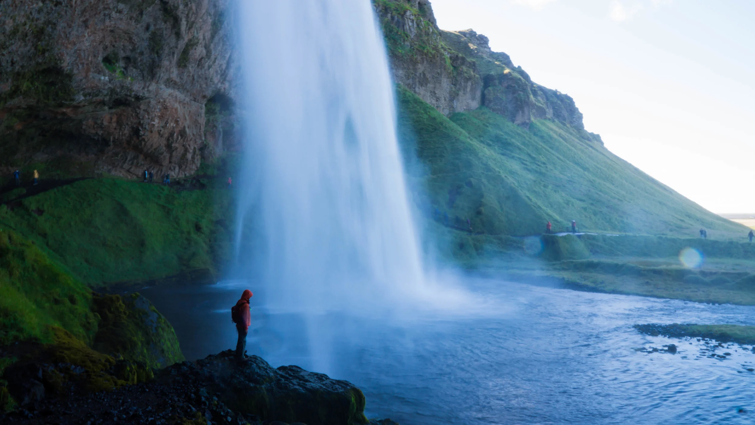 A waterfall in a lush green landscape with several people standing on a trail near the water.
