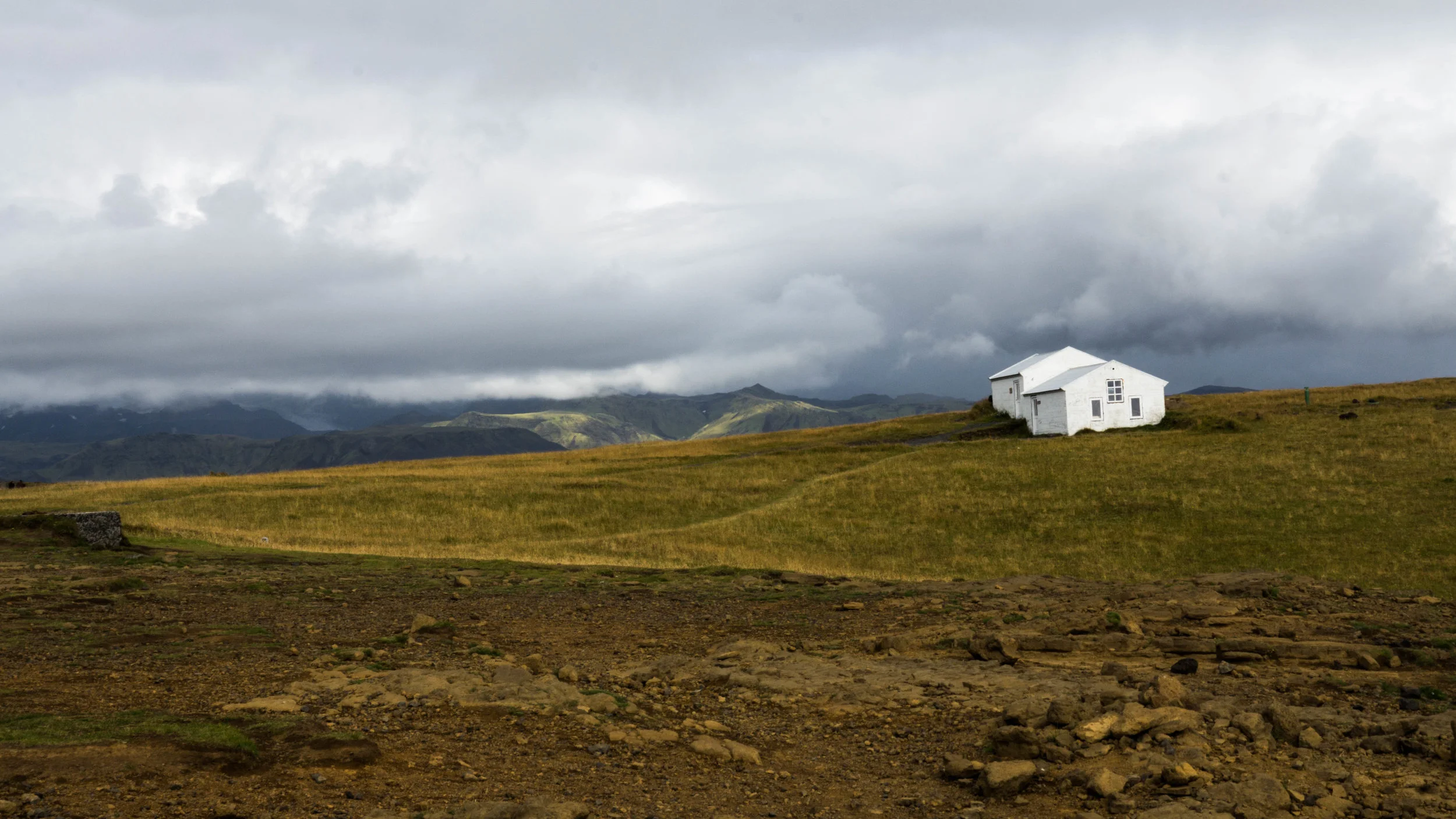 A white house with multiple sections and sloped roofs is situated on a grassy hillside, with mountains and a cloudy sky in the background.