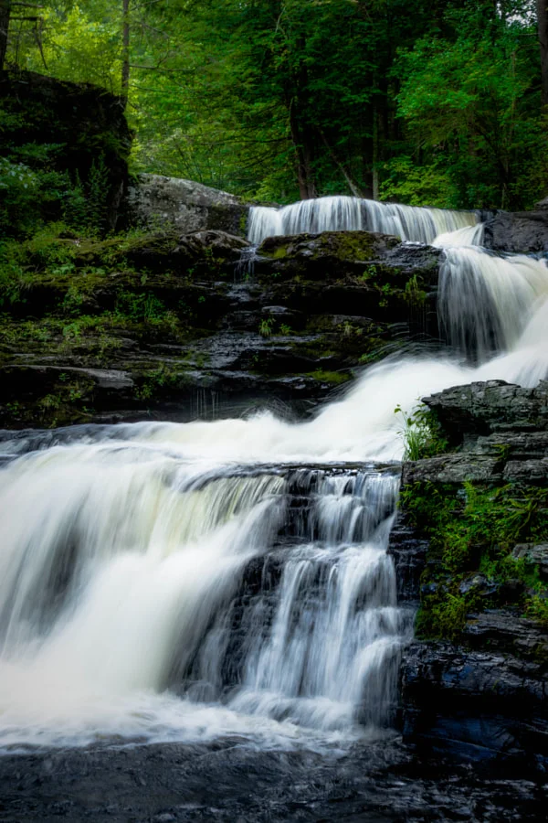 A multi-tiered waterfall flowing over rocks in a lush green forest.
