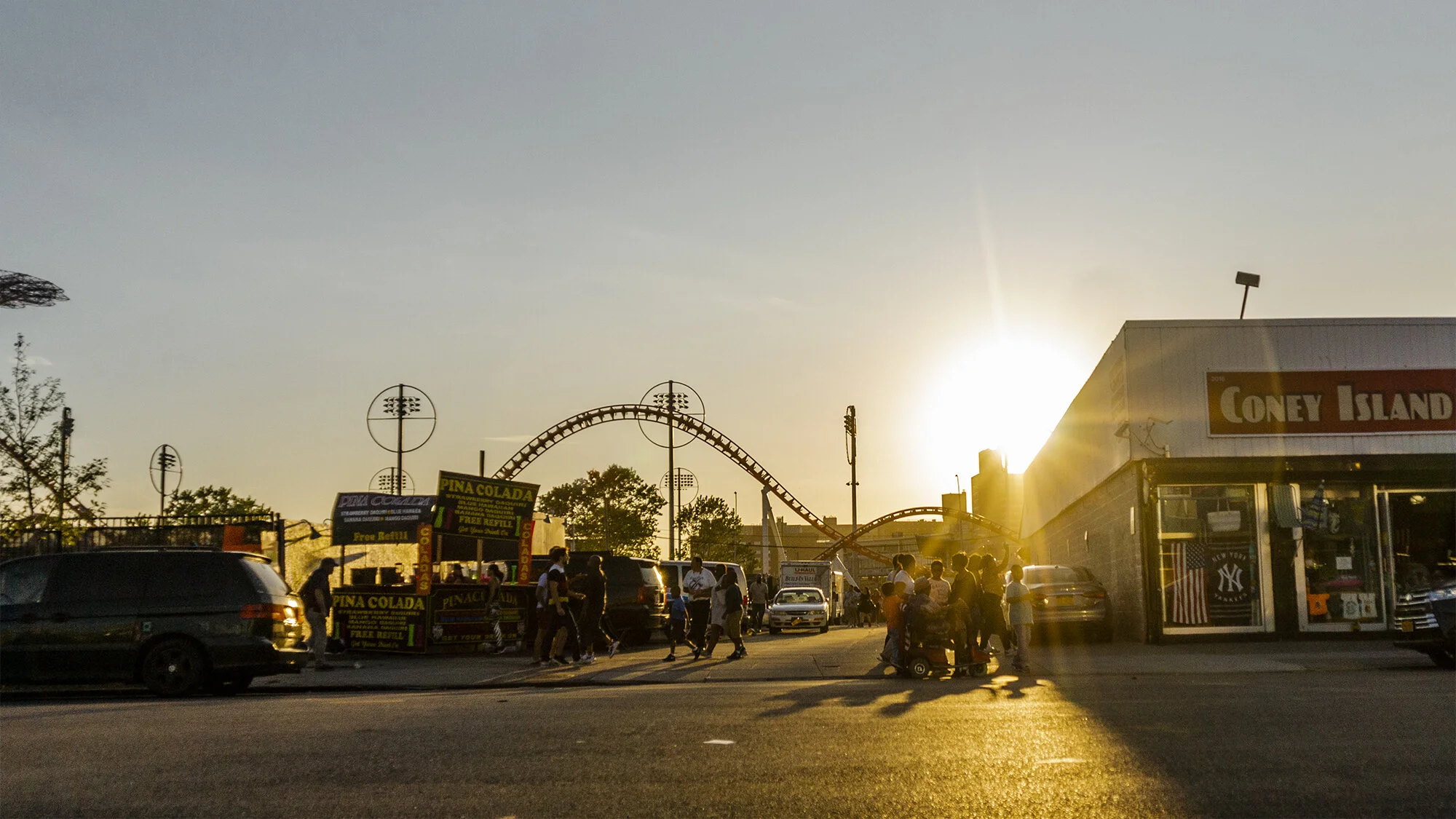 Sunset over an amusement park with a roller coaster, a food stand called Piña Colada, and a crowd of people walking and standing outside.