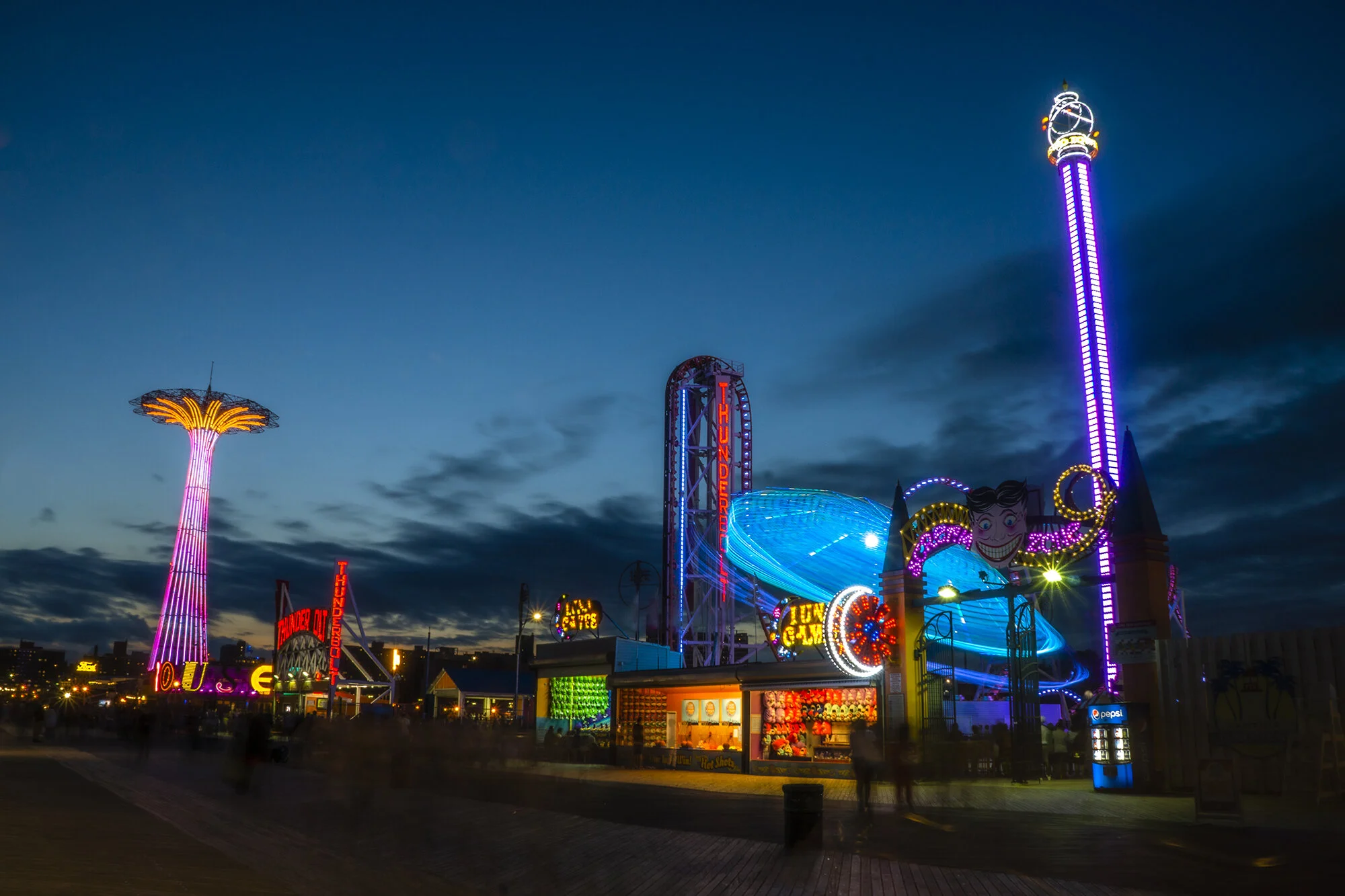 Colorful amusement park rides illuminated with neon lights at dusk, including a tall tower ride and a swing ride, with various game stalls and signage in the background.