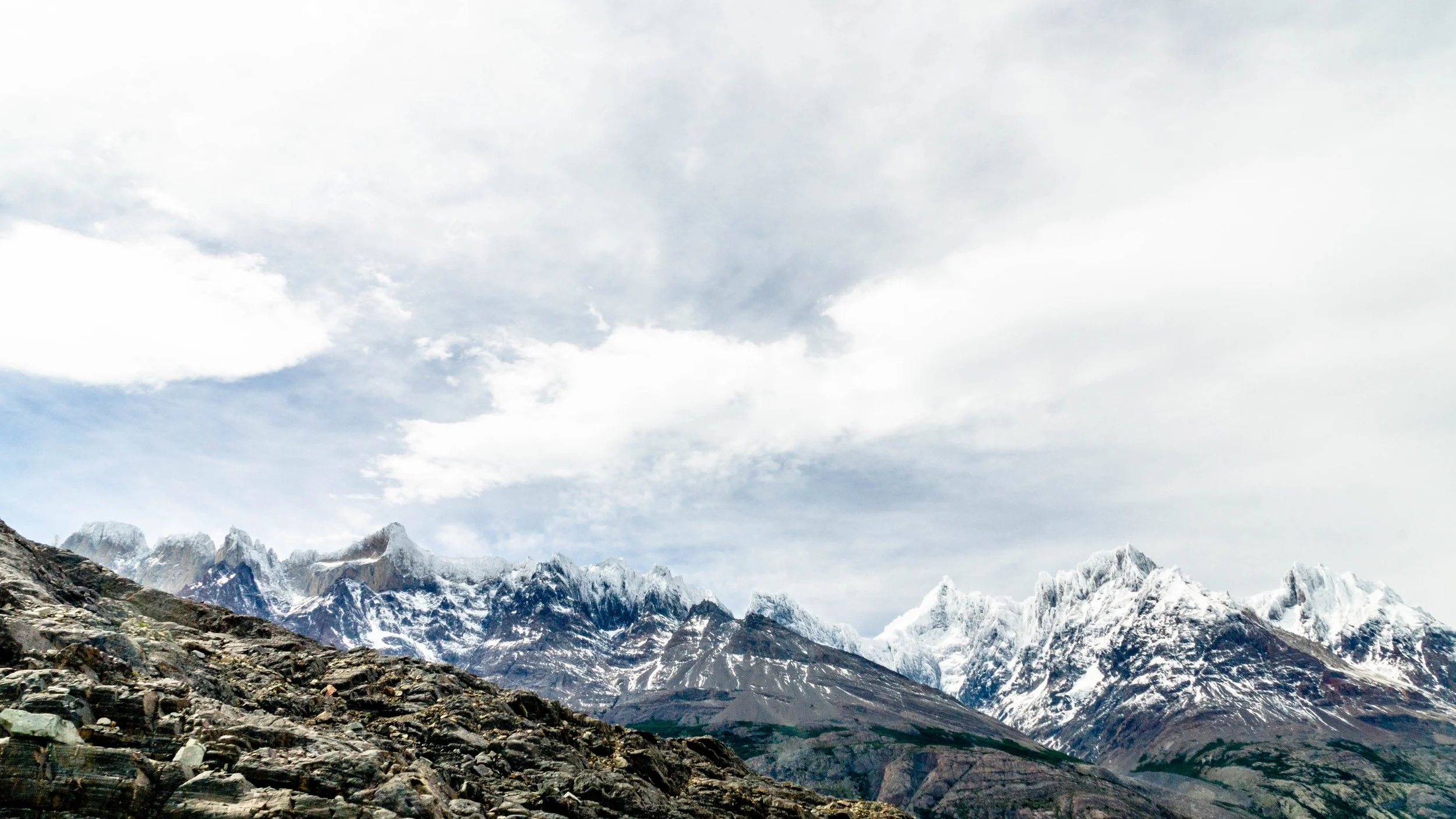 Snow-capped mountains under a cloudy sky with rocky terrain in the foreground.