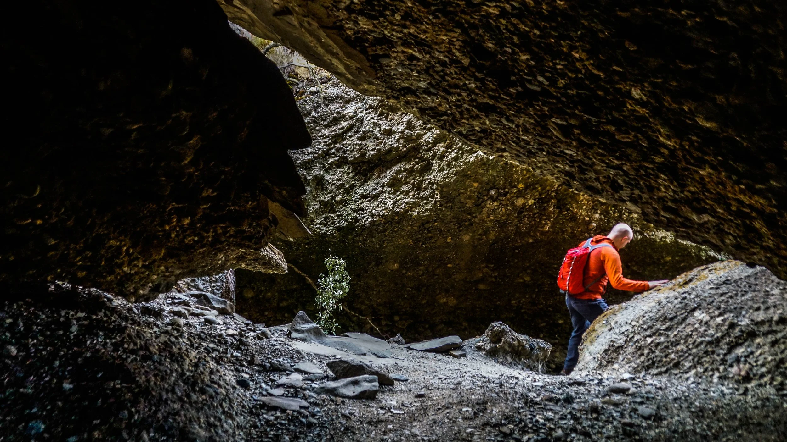 A person with a red backpack and orange jacket exploring a rocky cave or crevice.