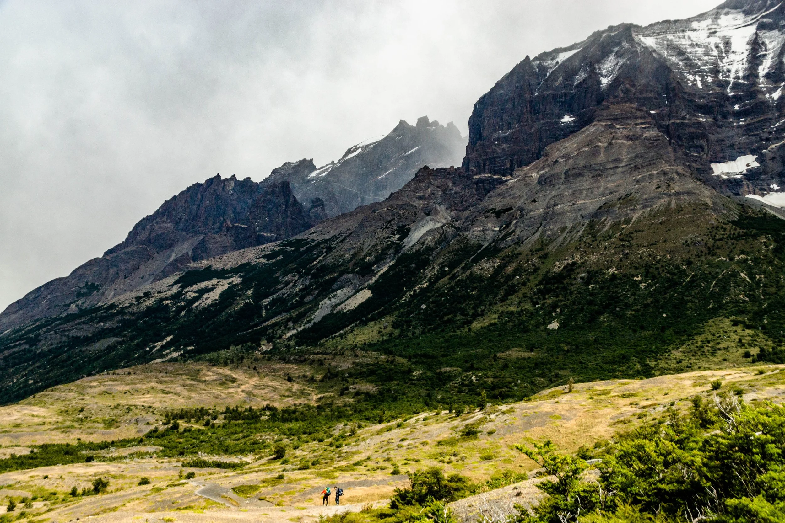 Two hikers walking on a trail in a vast mountain landscape with rugged peaks and some snow patches under cloudy skies.