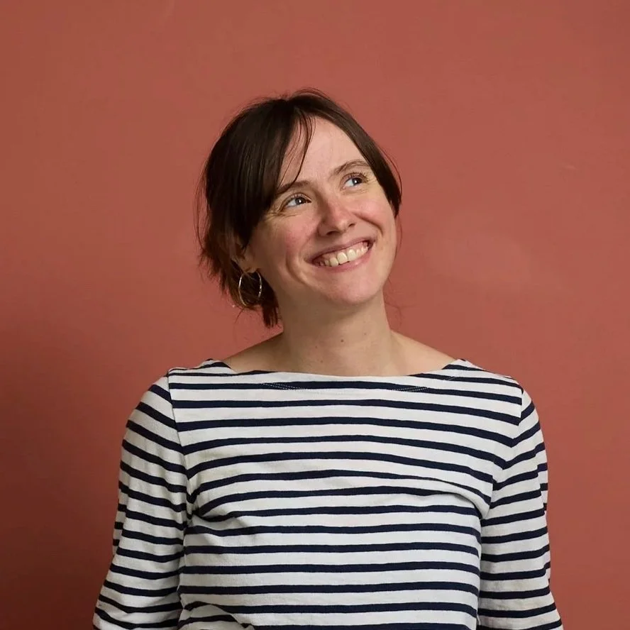 A smiling woman with short brown hair wearing hoop earrings and a black-and-white striped shirt, standing against a pink wall.