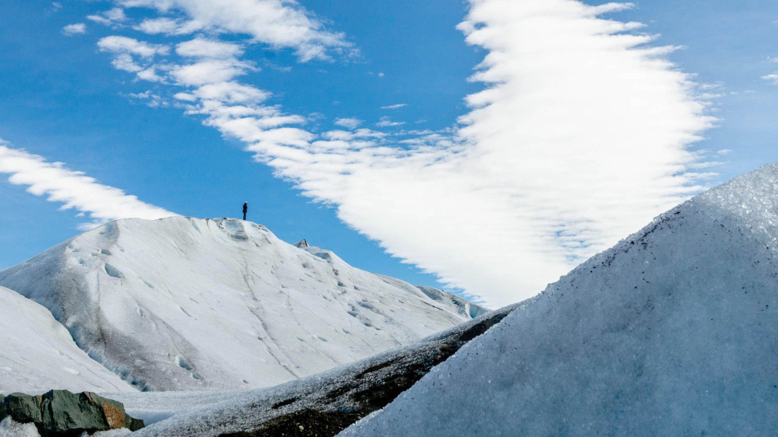 A person standing on a snowy mountain slope under a partly cloudy blue sky.