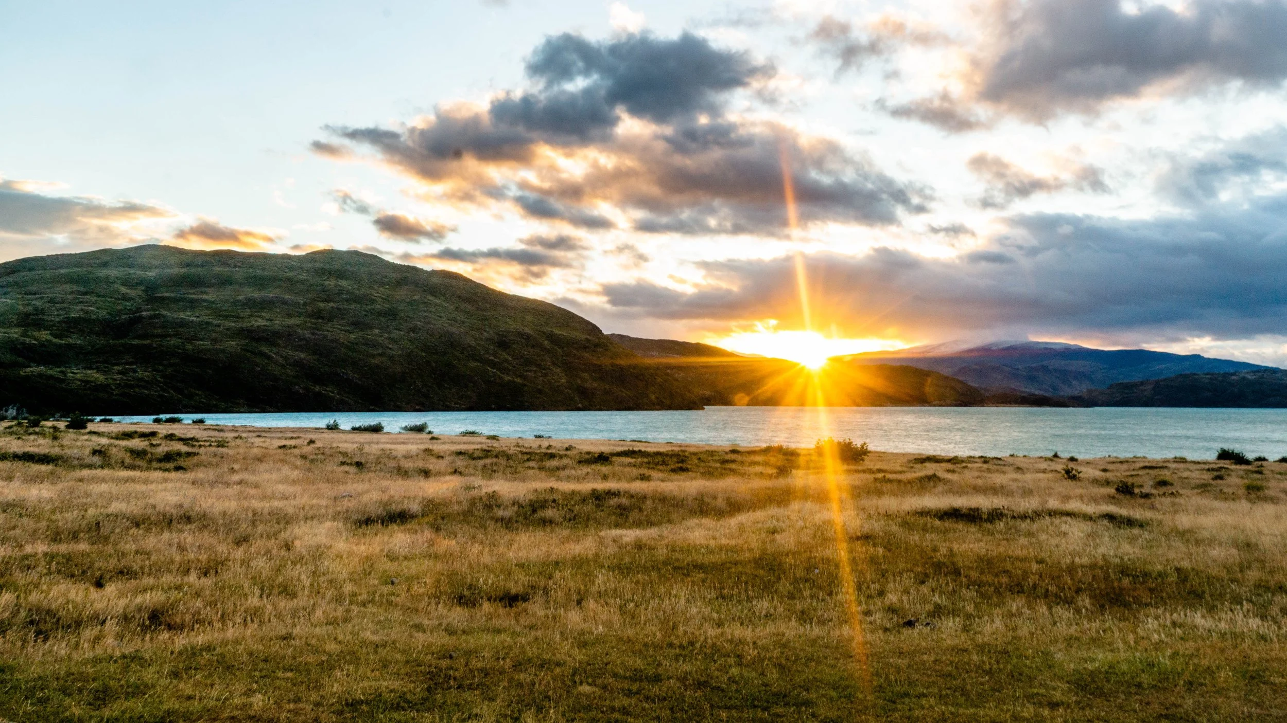A scenic landscape with a body of water reflecting the sunset, surrounded by rolling hills and mountains under a cloudy sky.