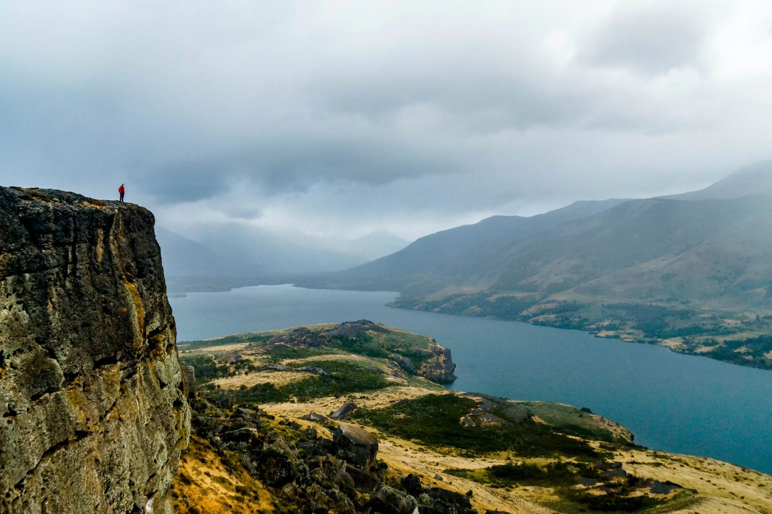 A person in an orange shirt standing on a rocky cliff overlooking a lake and mountains under a cloudy sky.