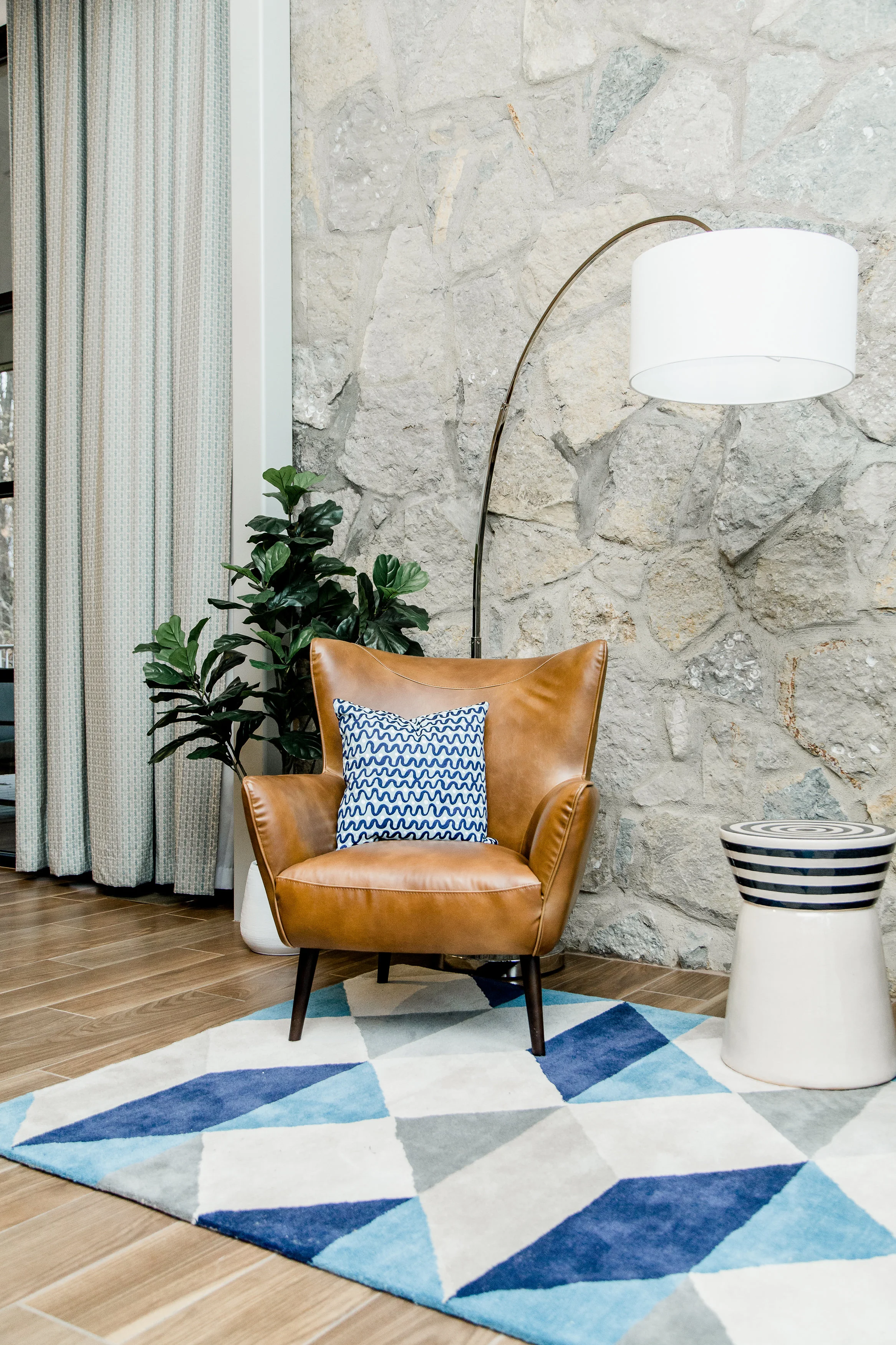 A note from the designer: Midcentury design can look too linear and stark, if you're not careful. So we used plants to warm up the space, a little. We were lucky that the client wanted to keep the original stone walls in the living room space, as well. That was a beautiful texture to play off of. We placed two beautiful leather chairs in front of the wall, in addition to a fiddle leaf fig plant. The chairs are a beautiful, supple leather which is in direct contrast with the rough, stone wall. And of course the fiddle leaf adds visual interest and dynamic movement.