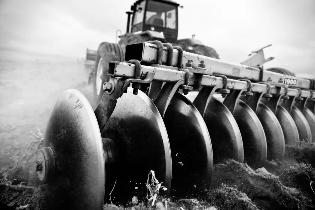 A black and white photo of a tractor plowing a field, with large rotating disks used for soil cultivation.