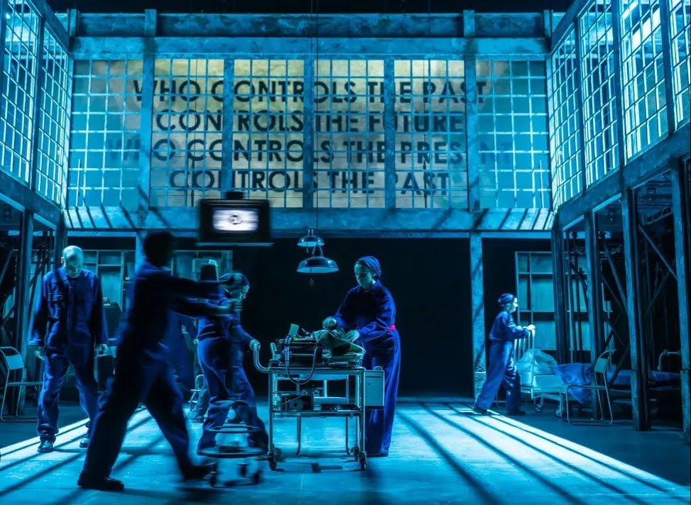 an image of a number of people, dressed the same, with heads down turned and dark lighting, under words on the wall saying "who controls the past controls the future. who controls the present controls the past"